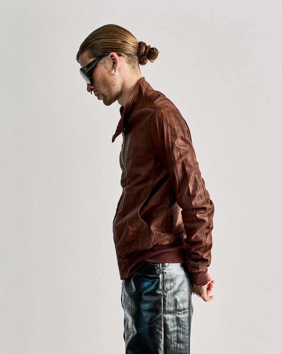A person with sunglasses and earbuds, wearing a brown leather Rick Owens jacket, stands sideways against a grey background with a reflective pose.