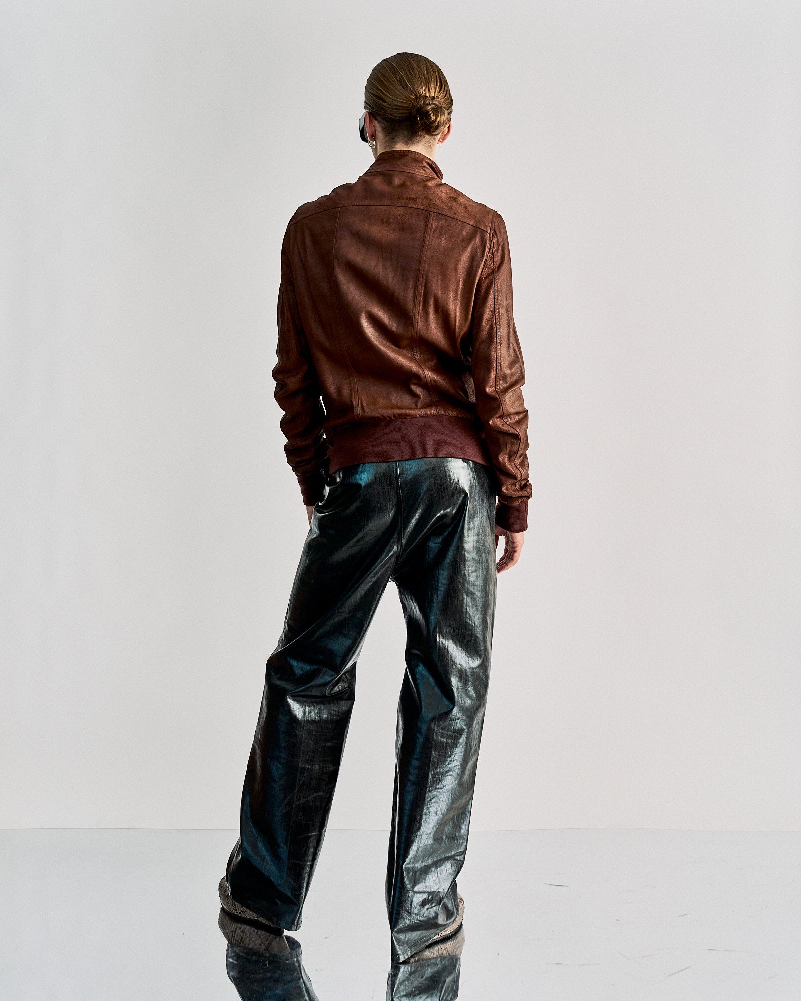 Person wearing a brown leather Rick Owens jacket and shiny black pants, standing with back to camera. The minimalist setting highlights a modern, edgy style.