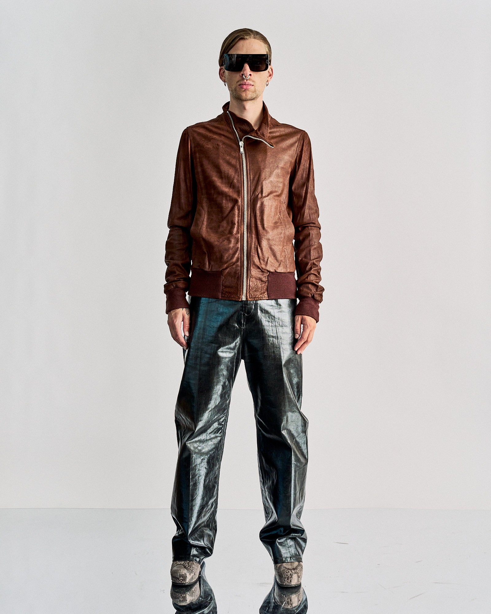 A person stands wearing a brown leather Rick Owens jacket, metallic silver pants, and snakeskin shoes against a plain background. 