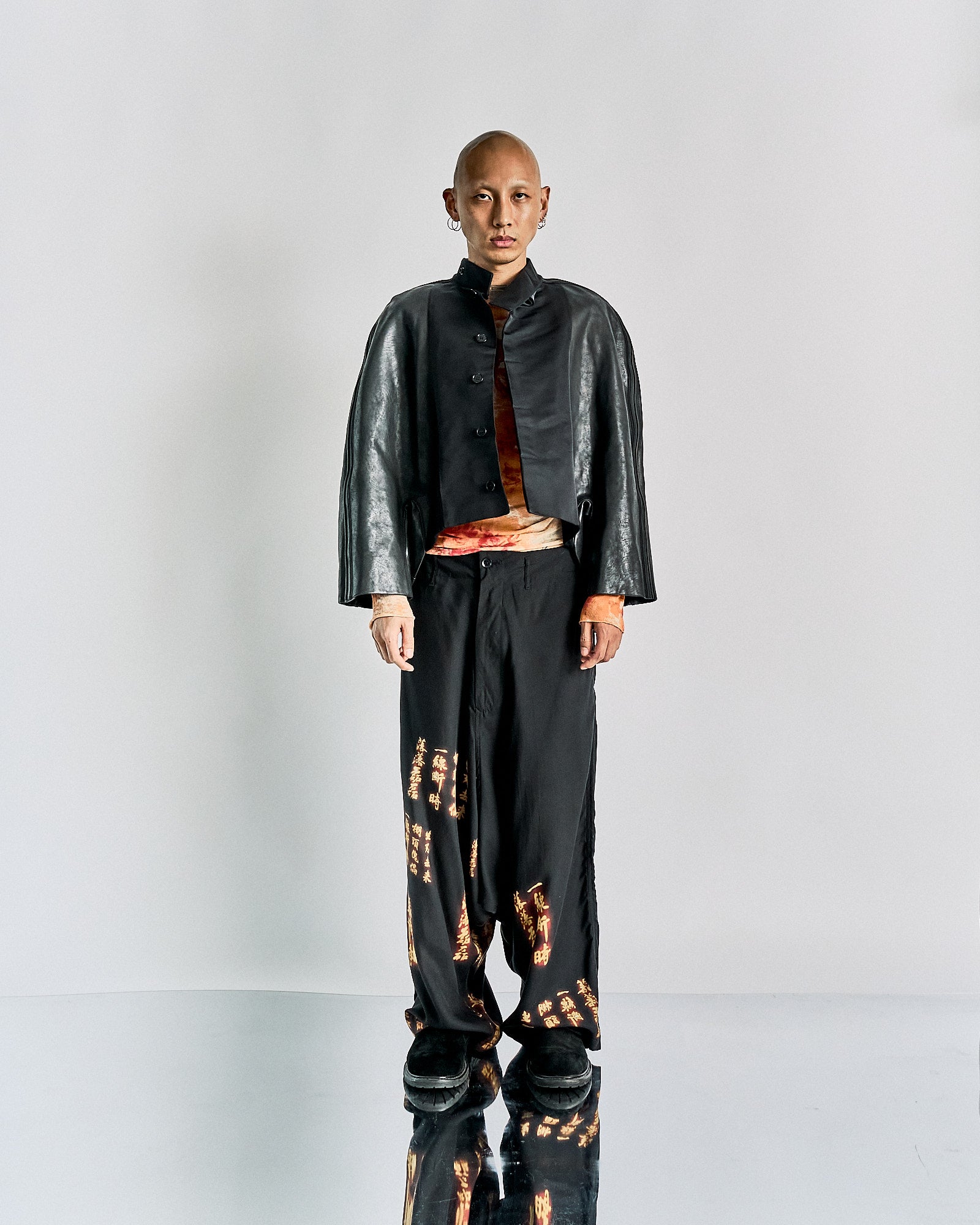 A person stands against a plain background, wearing a black leather Rick Owens jacket with an orange top beneath. Black pants feature orange patterns. 