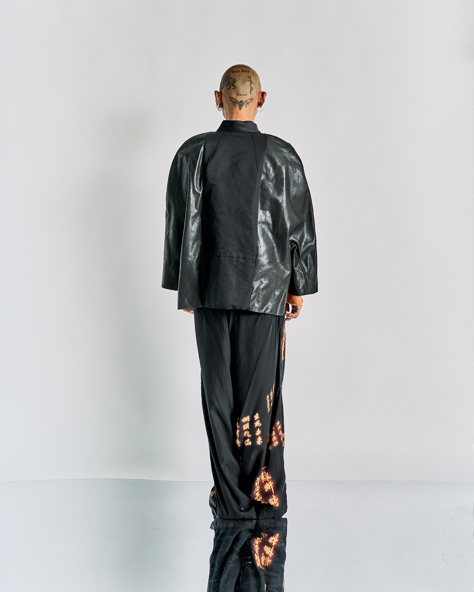 A person is standing with their back to the camera, wearing a black Rick Owens leather jacket and wide pants with orange patterns. The background is plain white.