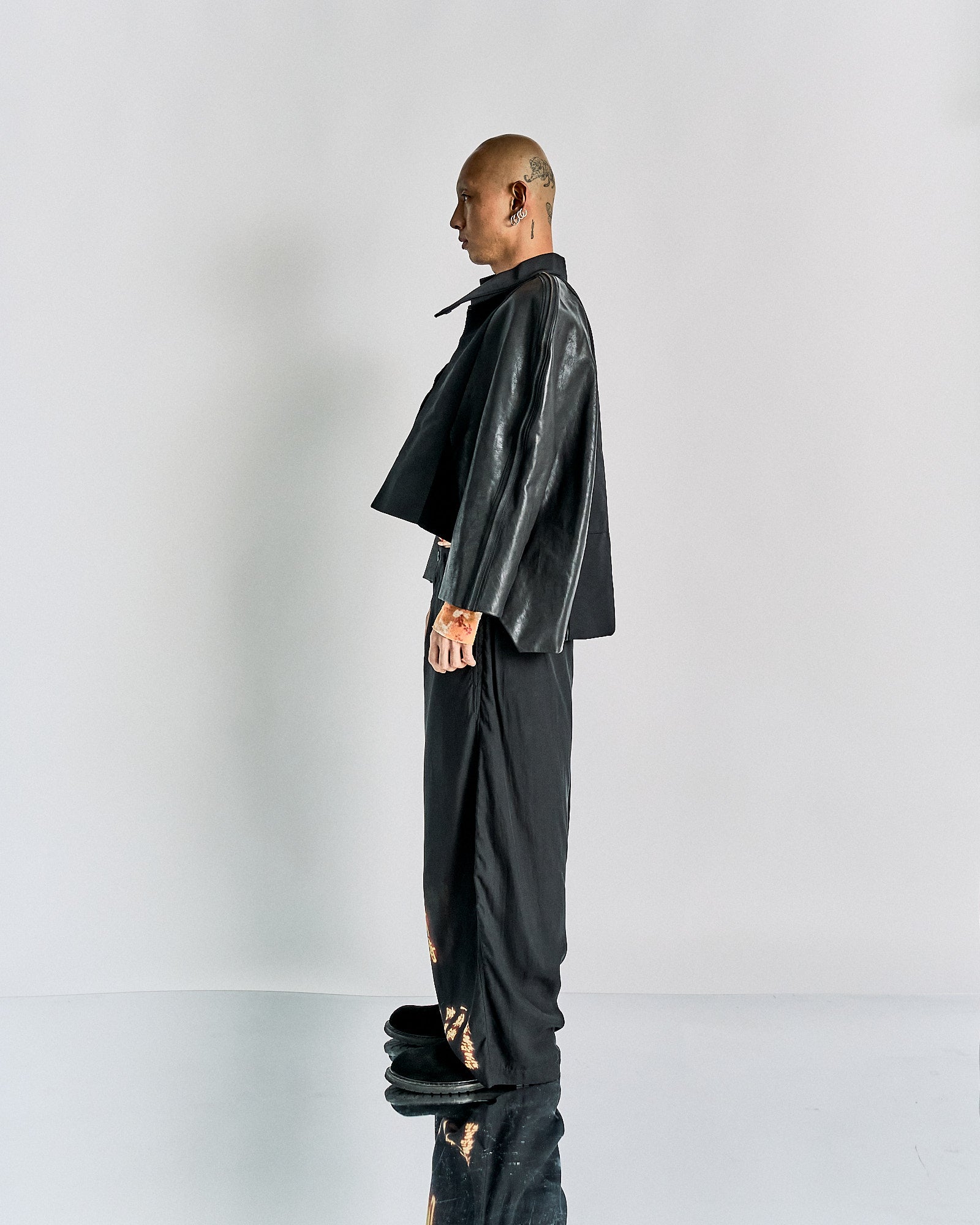 A side profile of a person standing against a plain background. They wear a black leather Rick Owens jacket and loose black pants, conveying a modern, edgy style.
