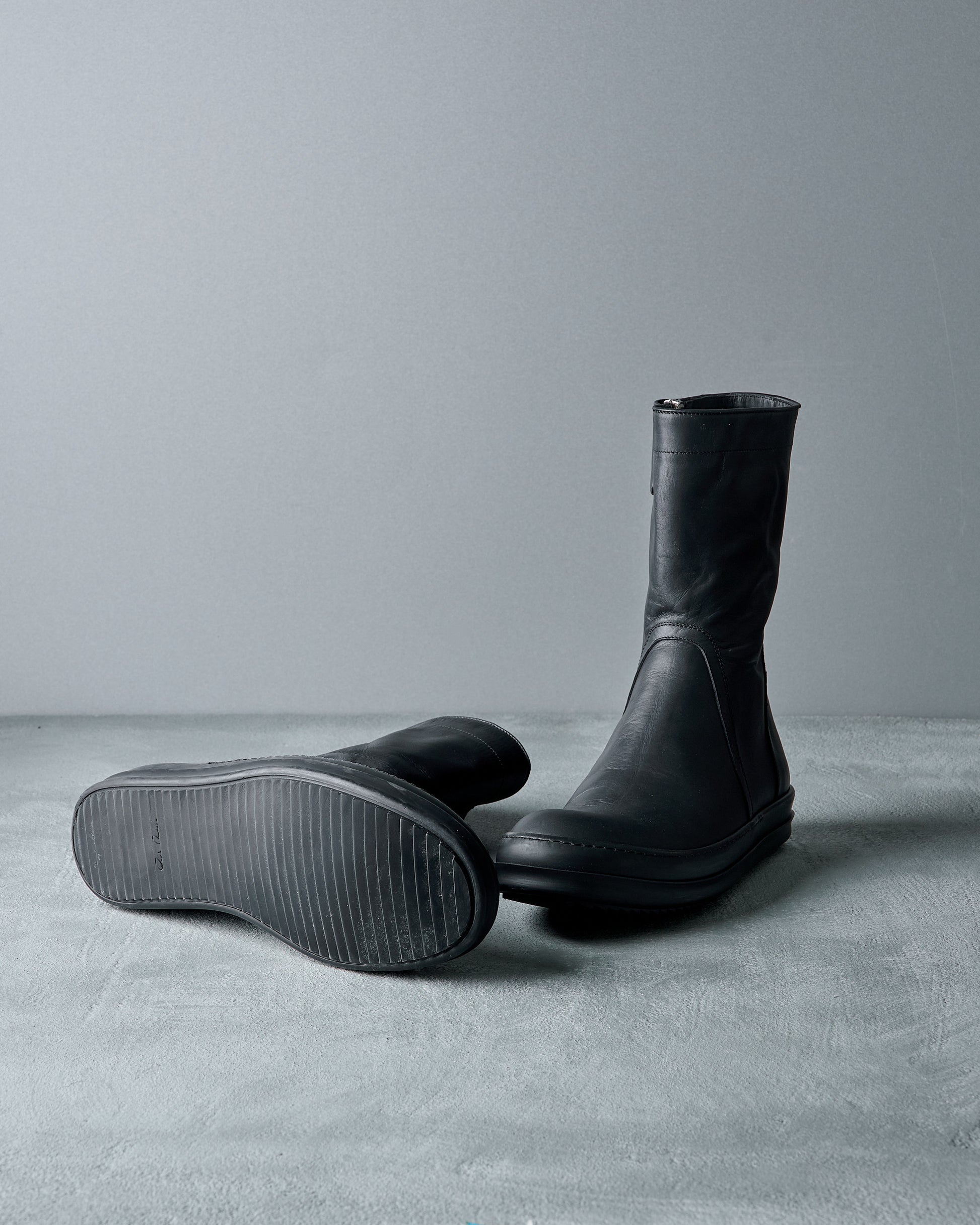 Pair of black rubber and leather Rick Owens boots on a gray surface with a neutral background