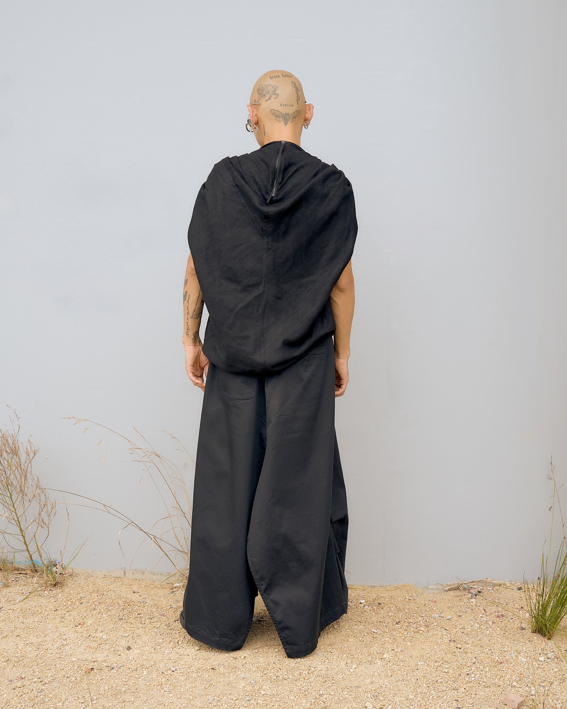 Bald person with tattoos stands facing a blank wall, wearing flowing black Rick Owens top and pants. Sandy ground with sparse grass creates a minimalist tone.