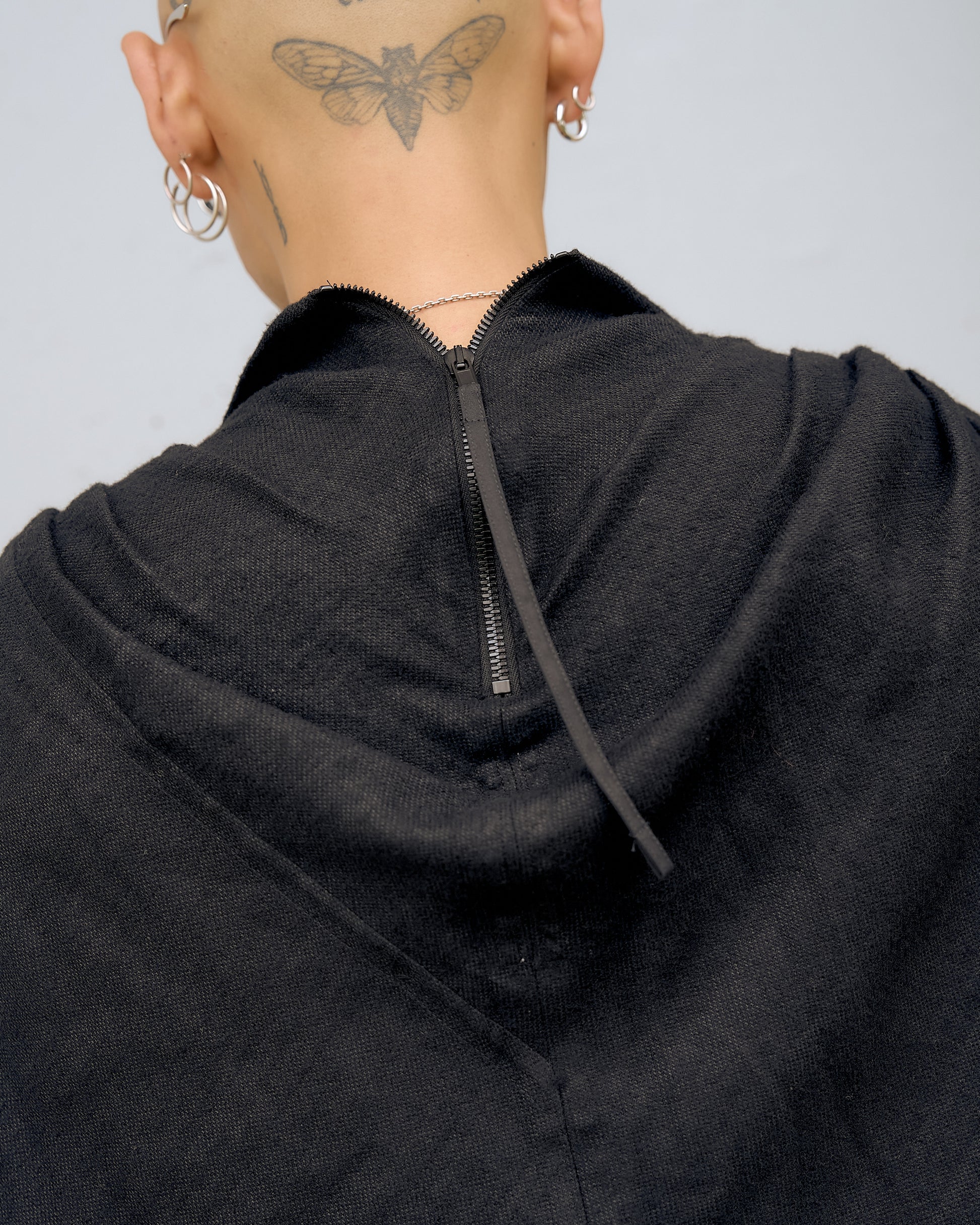 Close-up of the back of a person’s head with a butterfly tattoo, wearing silver earrings and a necklace. A black hooded Rick Owens garment is partially zipped.