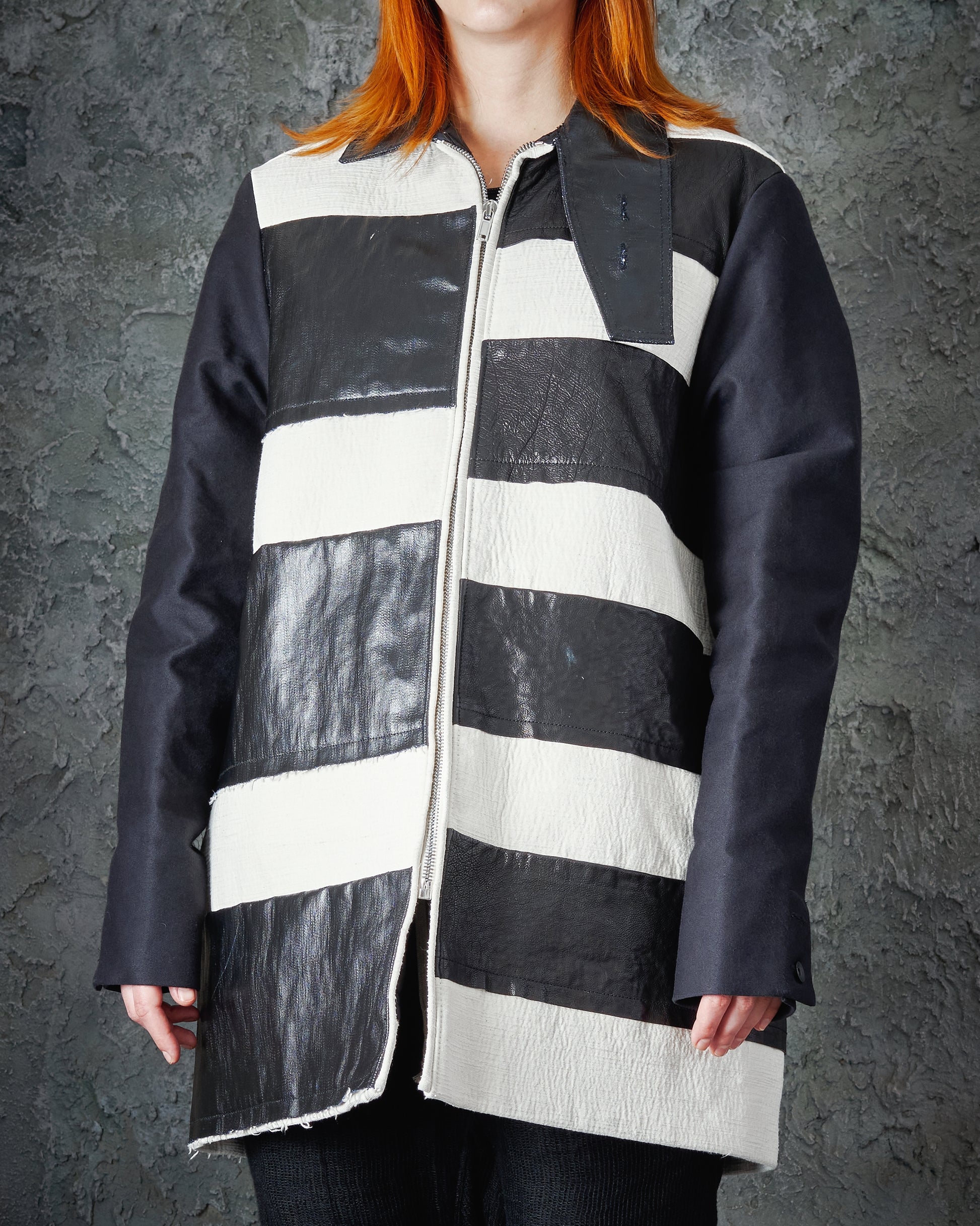 Person with red hair wearing a black and white striped leather Rick Owens jacket with dark sleeves. The textured grey background adds contrast.
