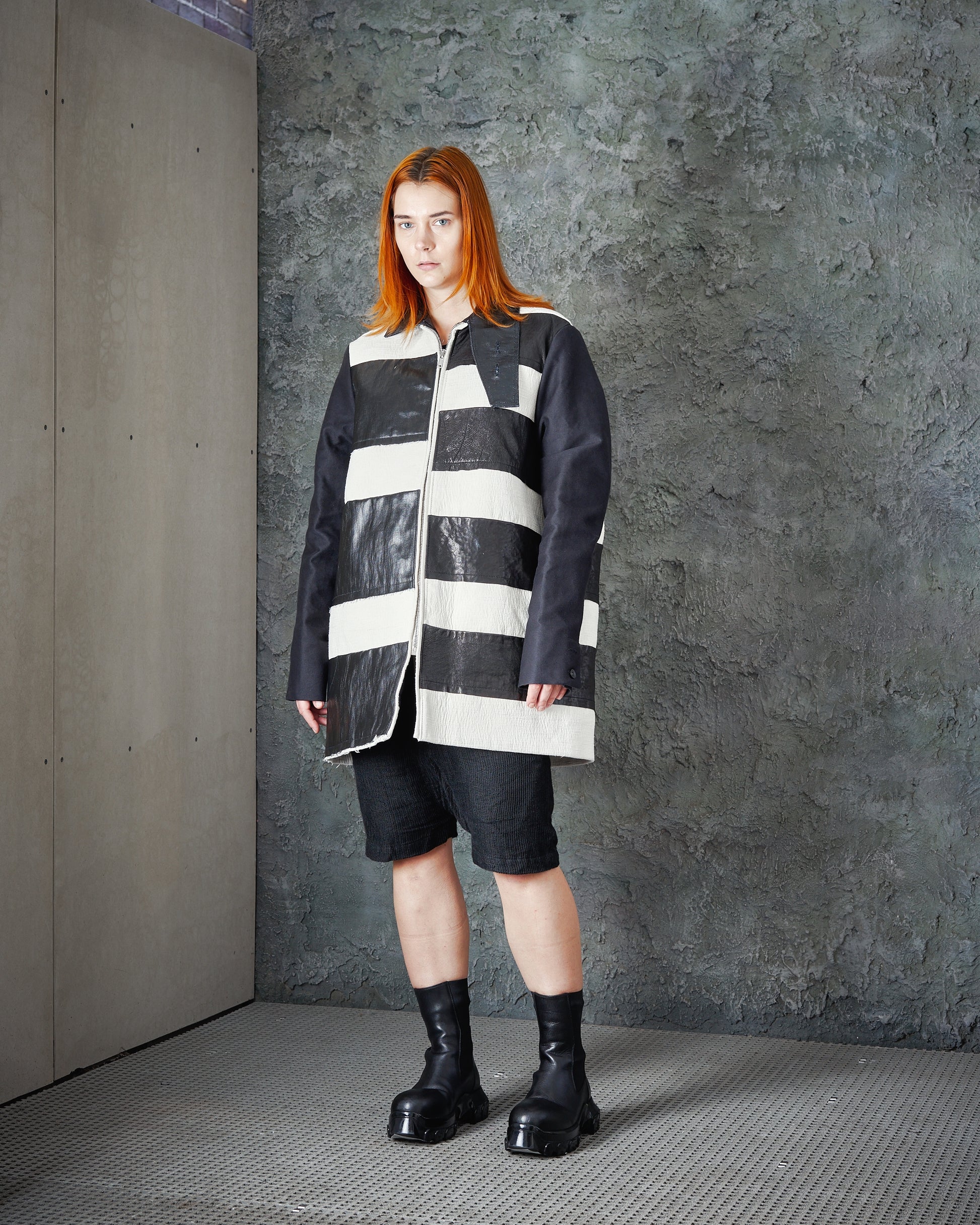 A person with red hair wearing a large black-and-white striped Rick Owens jacket, black shorts, and chunky black boots stands against a textured grey wall. 