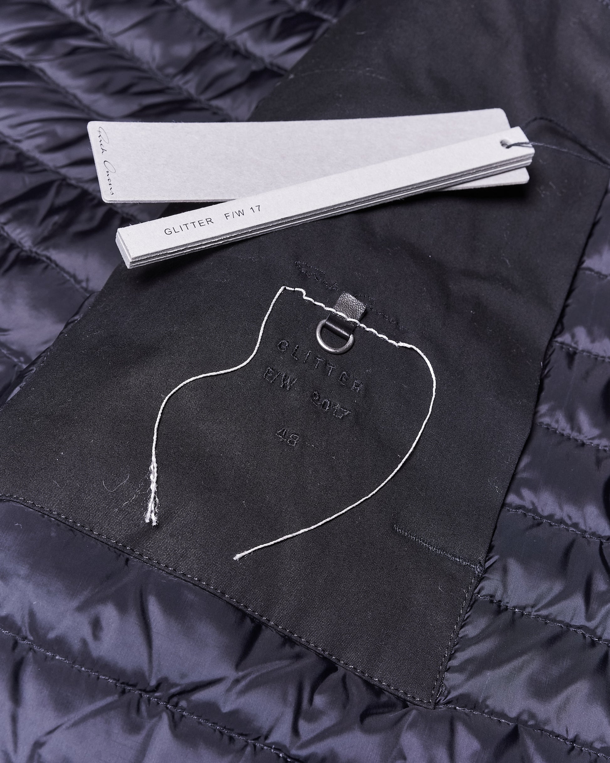 Close-up of Rick Owens garment elements featuring a black fabric patch with stitching and a hanging loop, placed over a blue quilted material. Nearby, paper tags labeled "GLITTER F/W 17" suggest fashion design context.