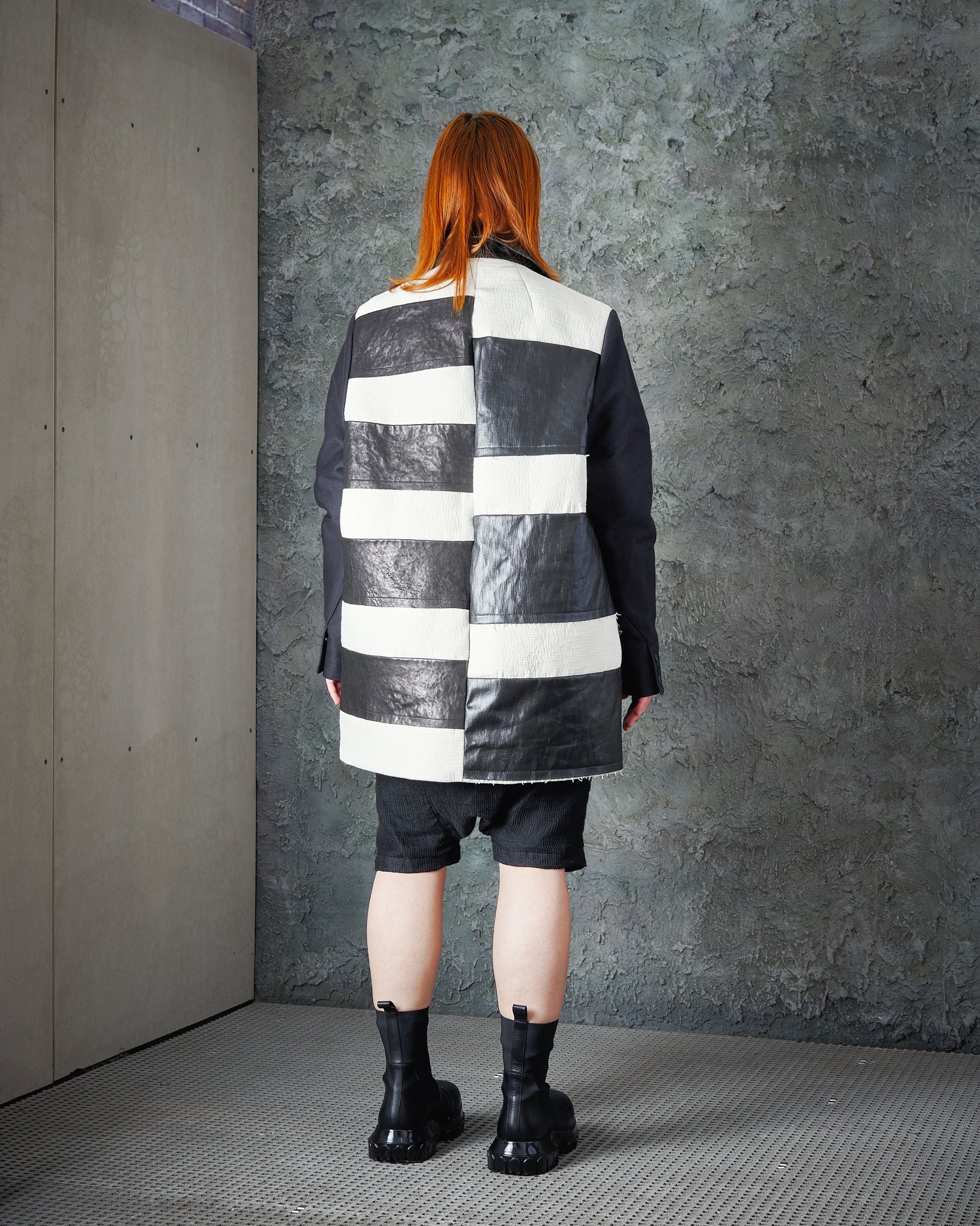 Person with long red hair stands facing a textured grey wall, wearing a black and white striped Rick Owens coat, black shorts, and chunky black boots. 