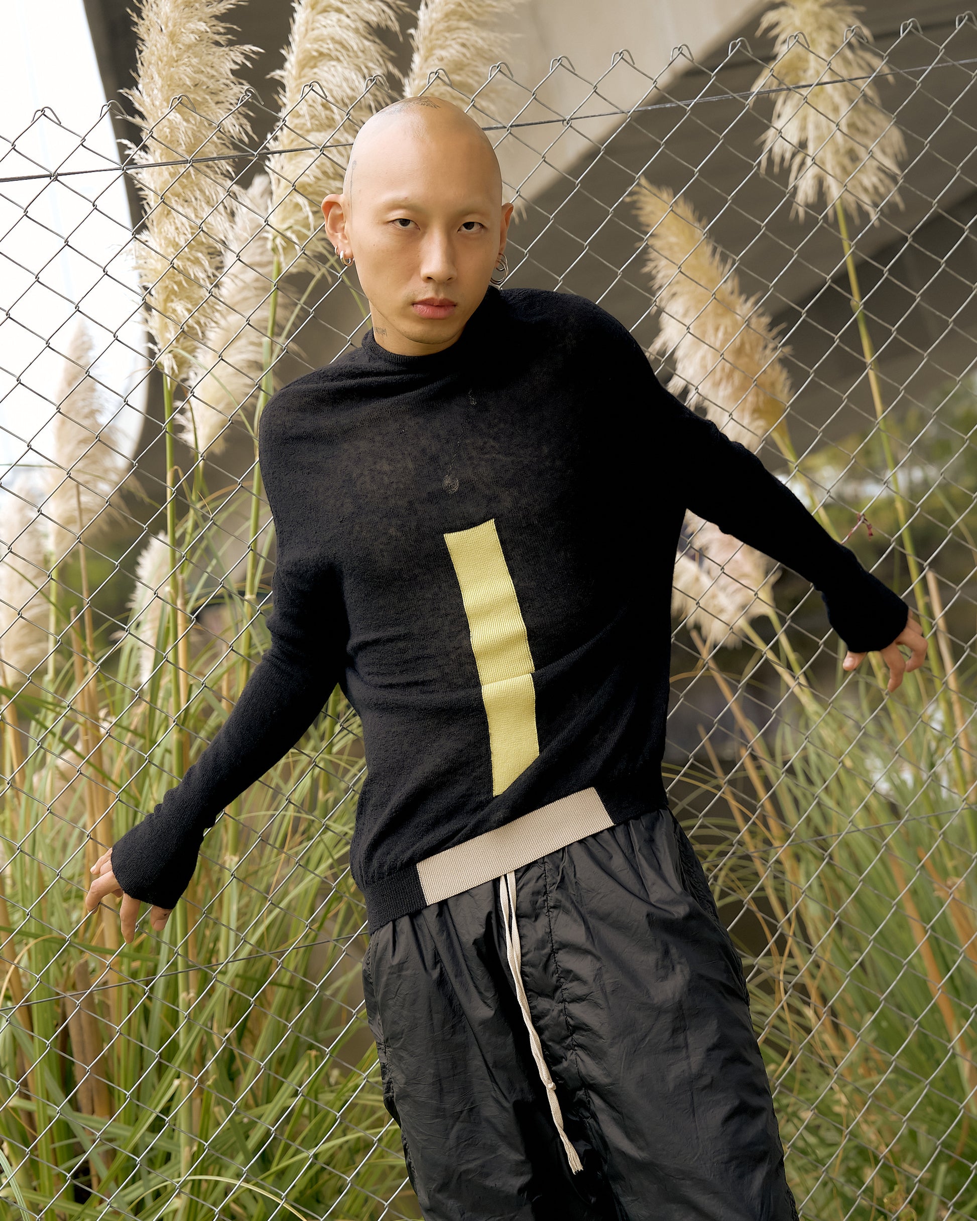 A bald individual poses confidently against a chain-link fence, wearing a black sweater with a yellow stripe and dark pants. Tall grasses in the background convey a natural setting.