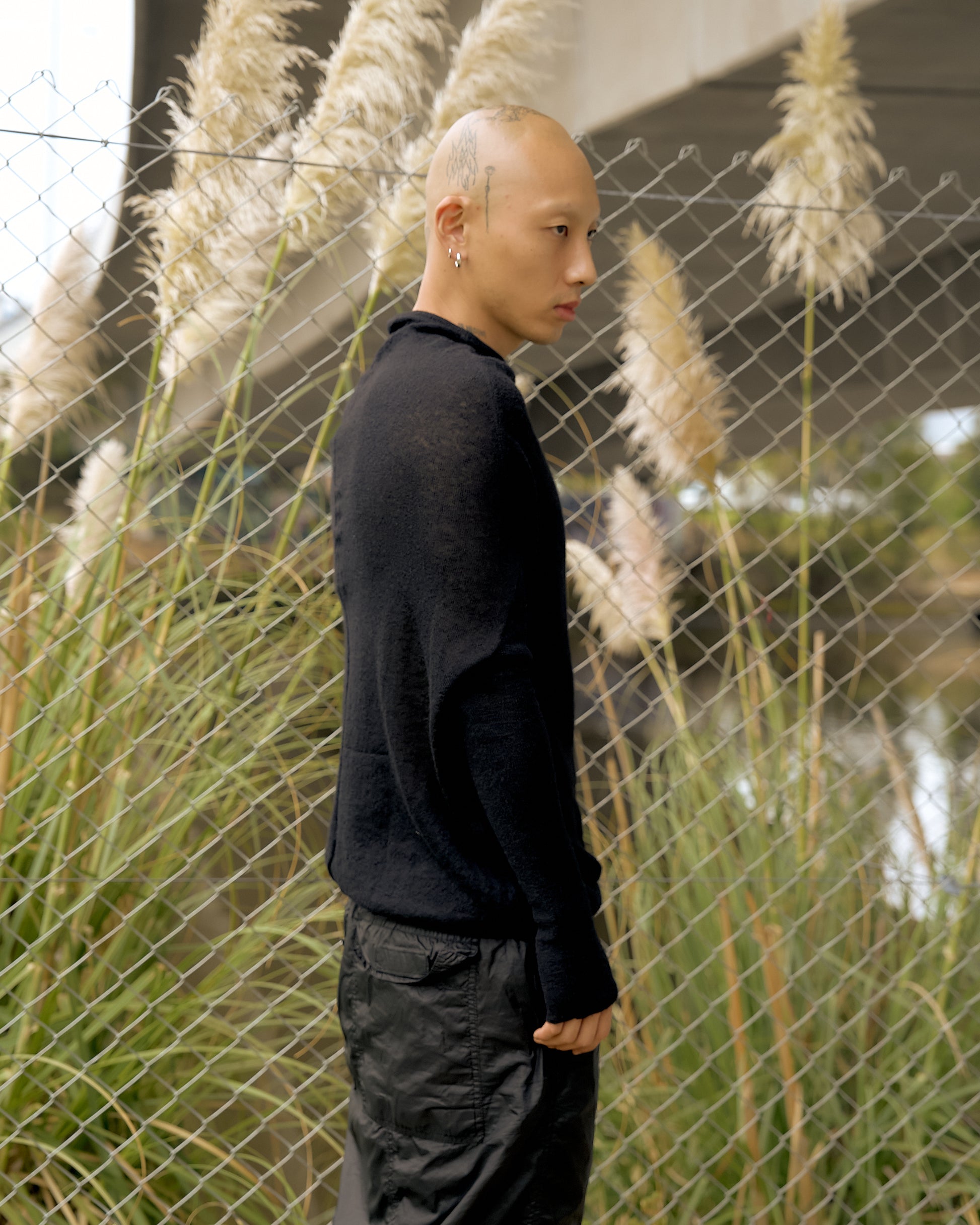 A person stands sideways in front of a chain-link fence adorned with tall grasses and pampas plumes. Wearing a black Rick Owens outfit, they convey a contemplative mood.