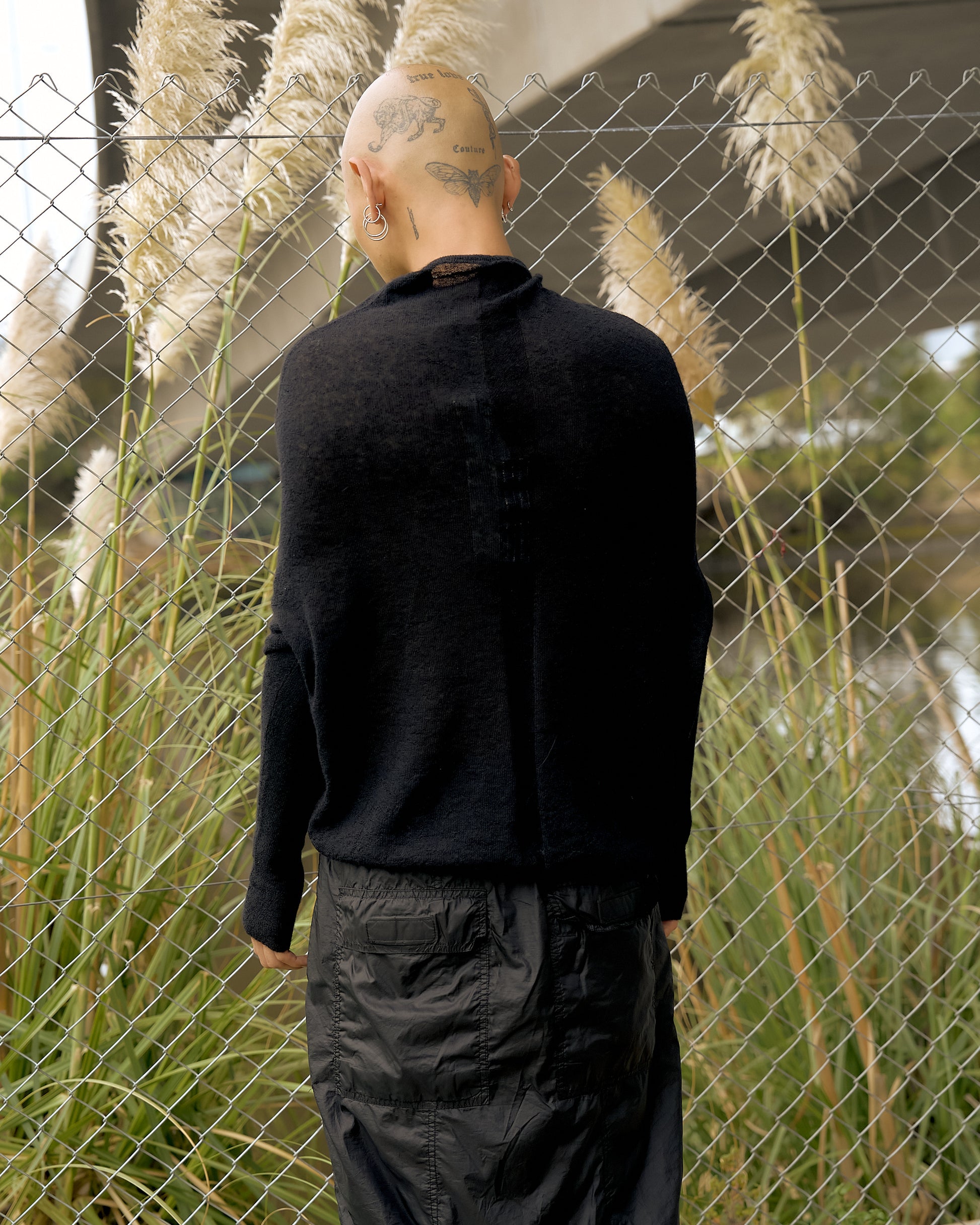A person with a shaved head and tattoos stands with their back to the camera, wearing a black Rick Owens sweater and pants. They stand in front of a chain-link fence with tall grasses, conveying a contemplative mood.