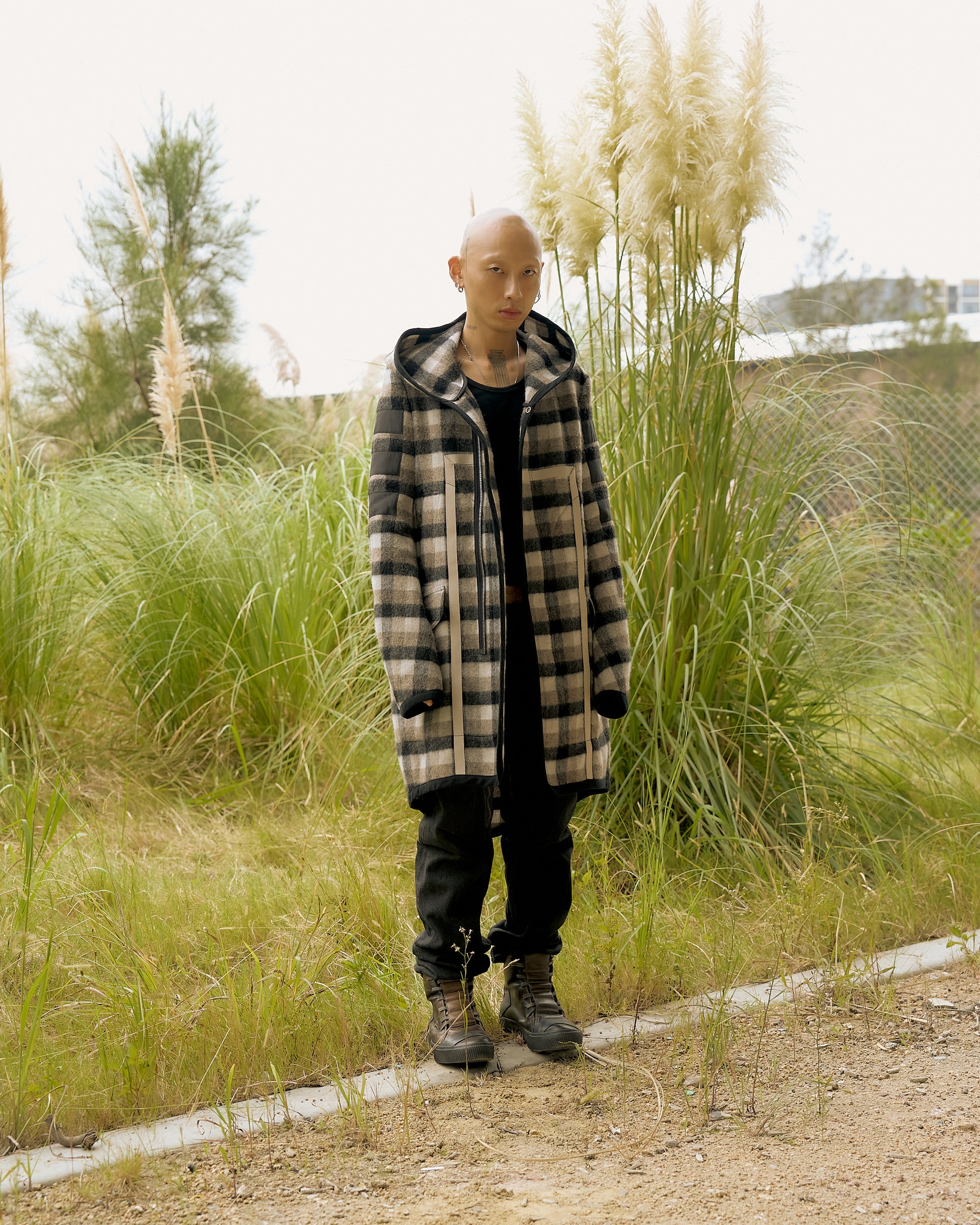 A person wearing a plaid Rick Owens coat and black outfit stands on grass near tall pampas plants. 