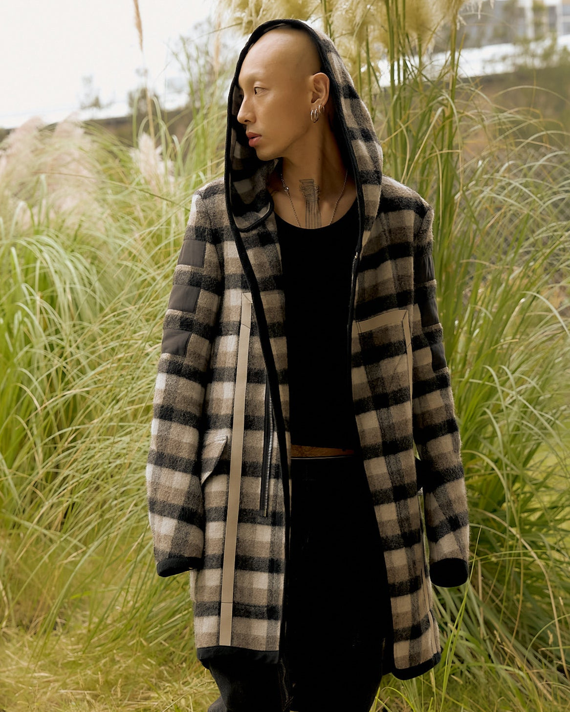 A person wearing a plaid Rick Owens coat with a hood stands in tall, lush grass, looking to the side.