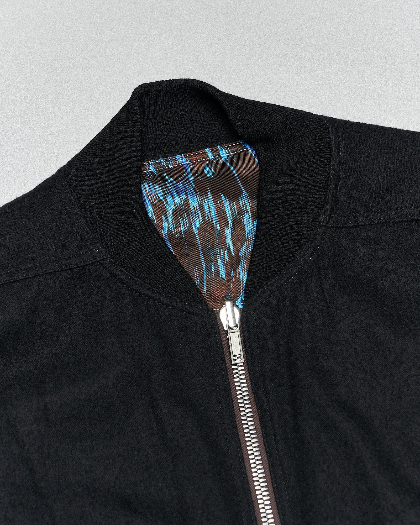 Close-up of a black Rick Owens jacket with a visible metallic zipper and collar, featuring a blue and brown patterned inner lining. The tone is modern and stylish.