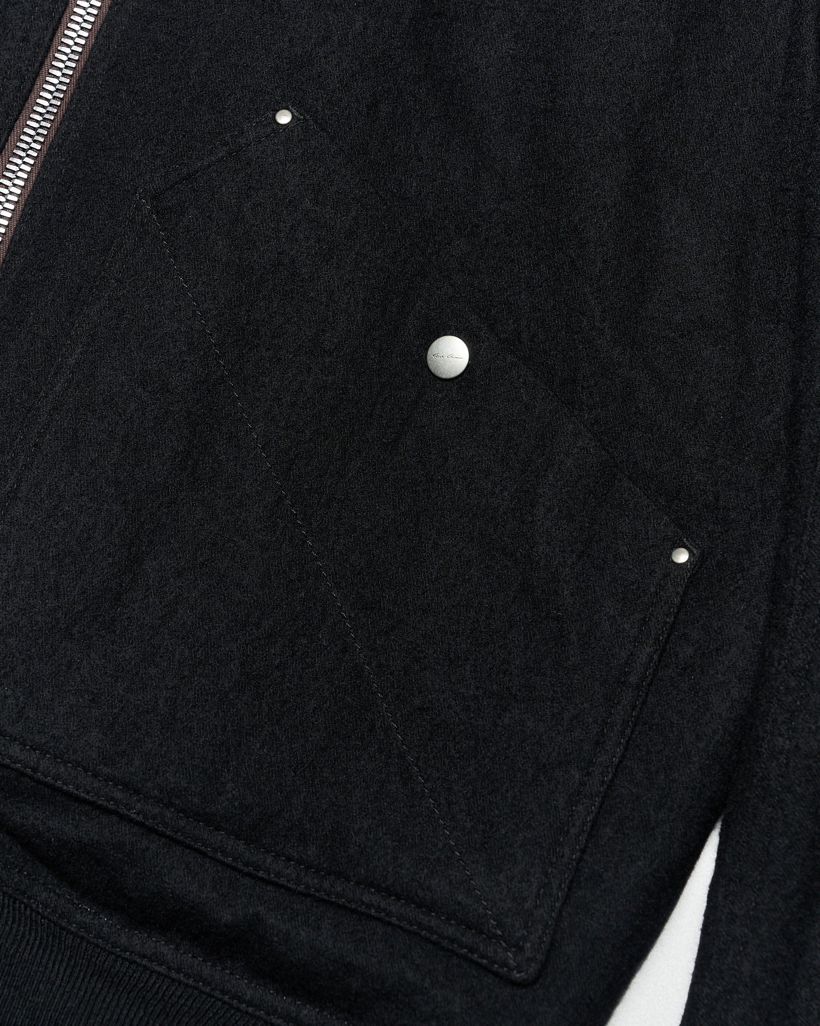 Close-up of a black textured fabric from a Rick Owens bomber jacket with a silver zipper running vertically. A pocket with metal studs is visible, conveying a sleek, modern style.