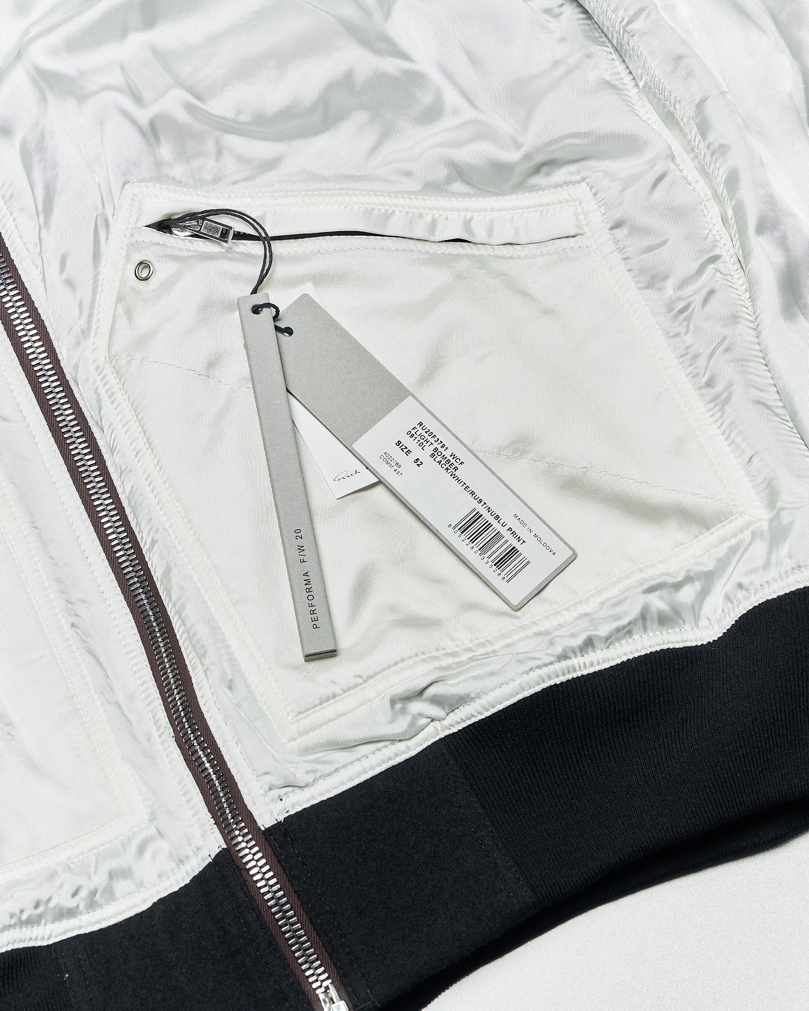 Inside of a Rick Owens  jacket with a white lining and a sewn-in zipper pocket. A grey tag with product details is attached, creating a clean, modern tone.