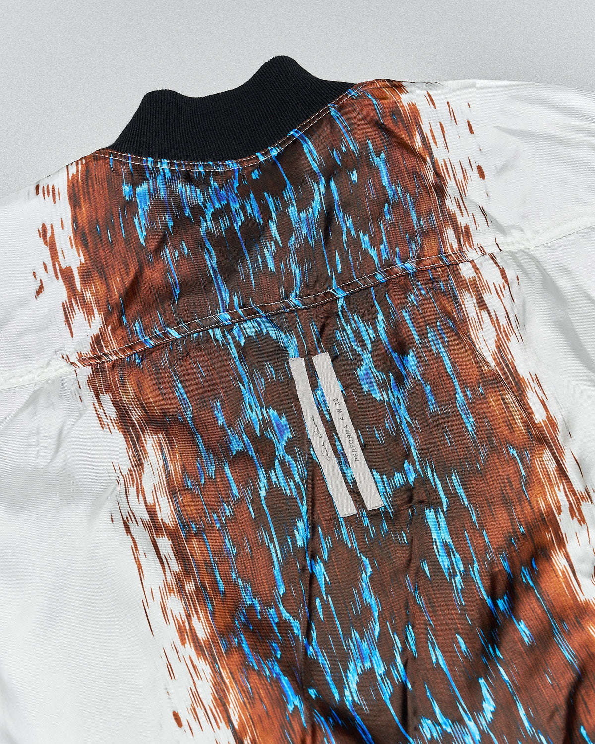 Person wearing a black zip-up Rick Owens jacket and shiny black pants stands on a reflective surface. The minimalist background creates a modern, edgy tone.