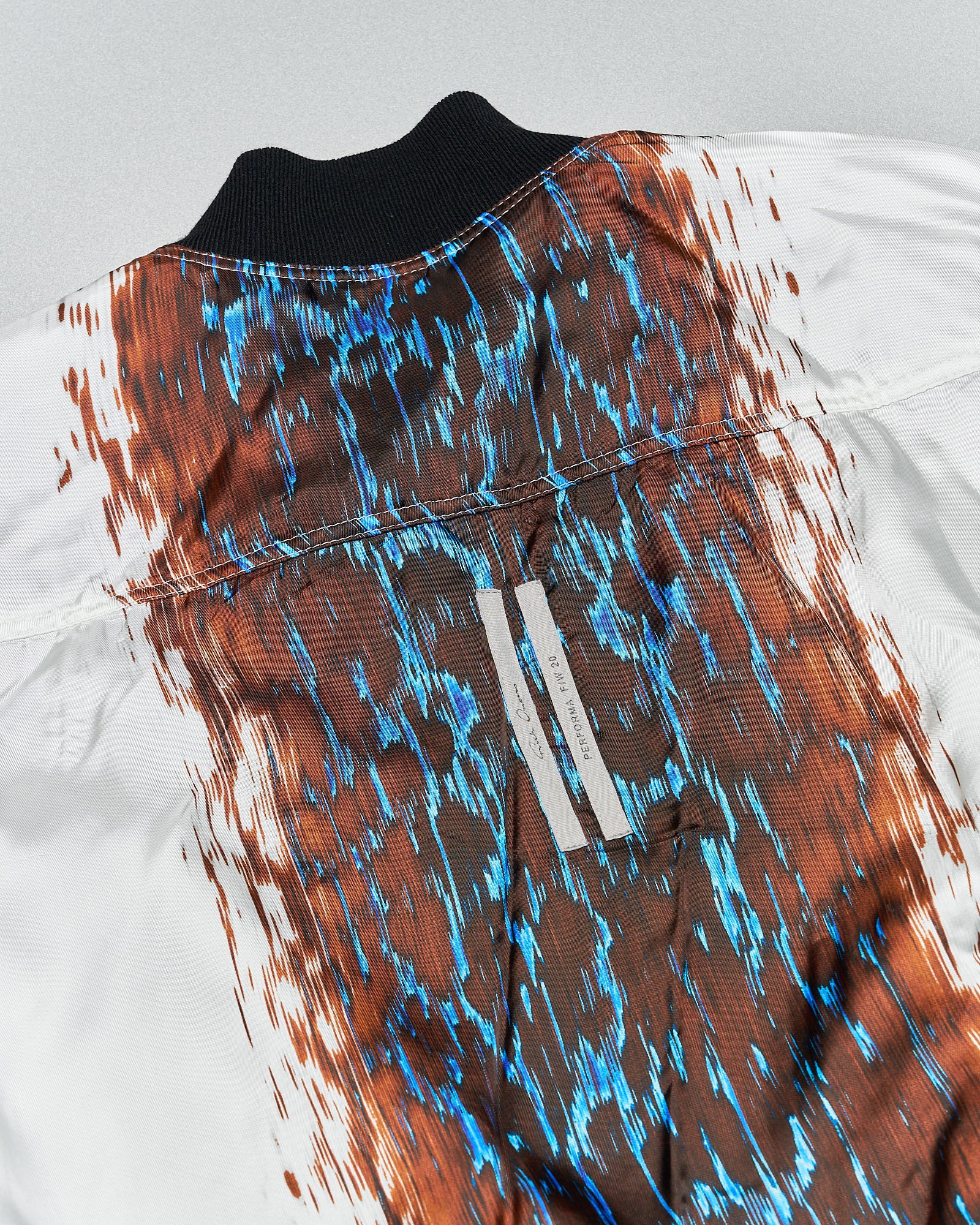 Close-up of a Rick Owens jacket's back showcasing an abstract pattern with vibrant blue and burnt orange streaks, over a white fabric. The texture appears dynamic.