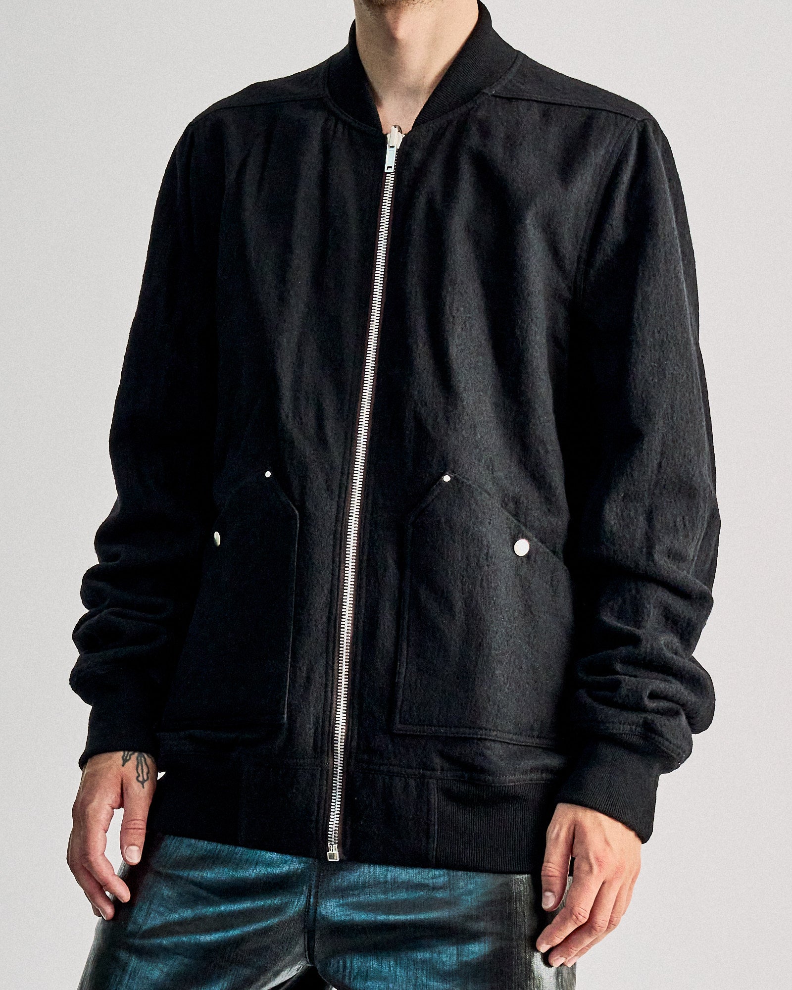 Person wearing a black zip-up Rick Owens jacket with silver buttons, paired with metallic pants. They stand against a plain light gray background.