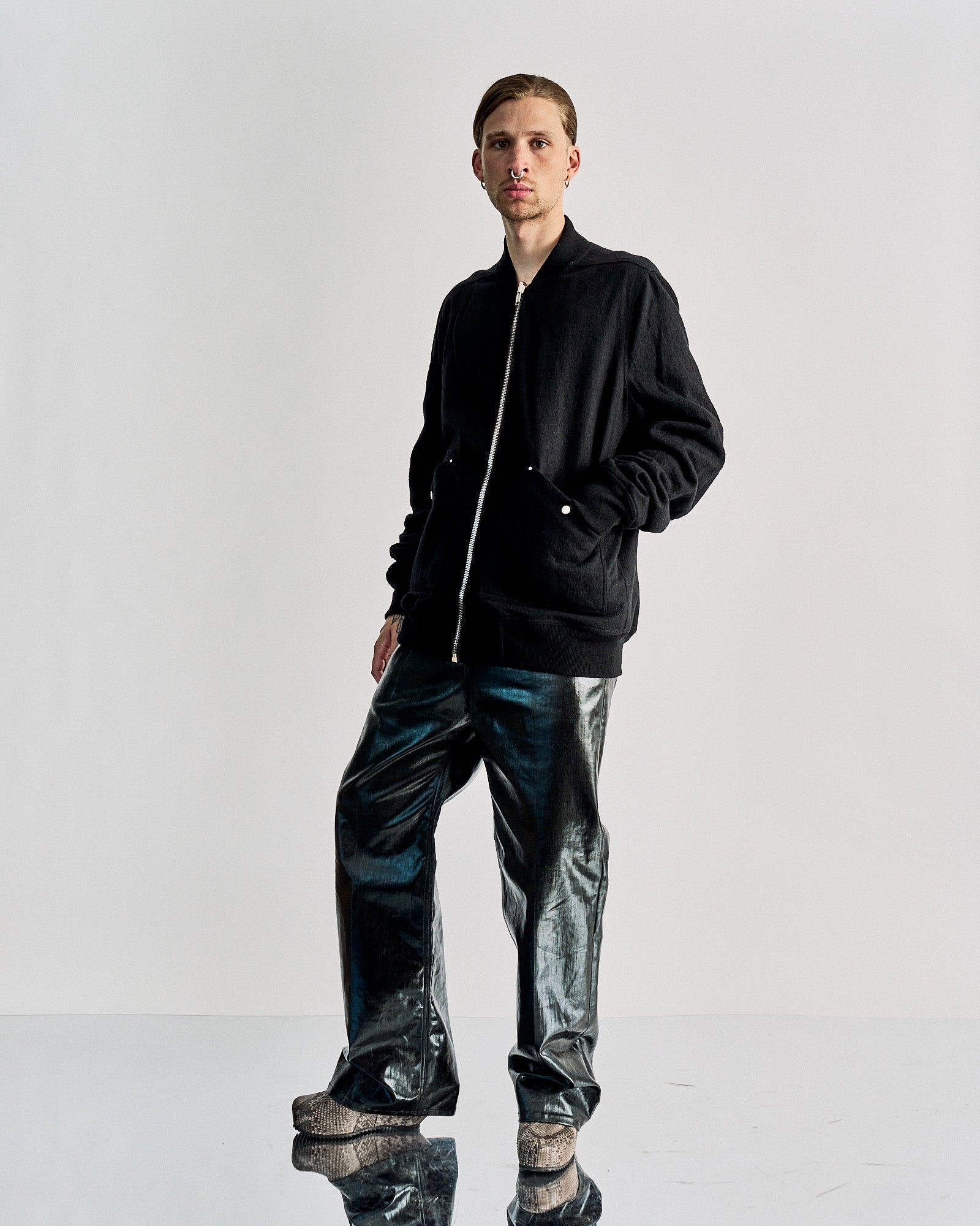 Person wearing a black zip-up Rick Owens jacket with silver buttons, paired with metallic pants. They stand against a plain light gray background.