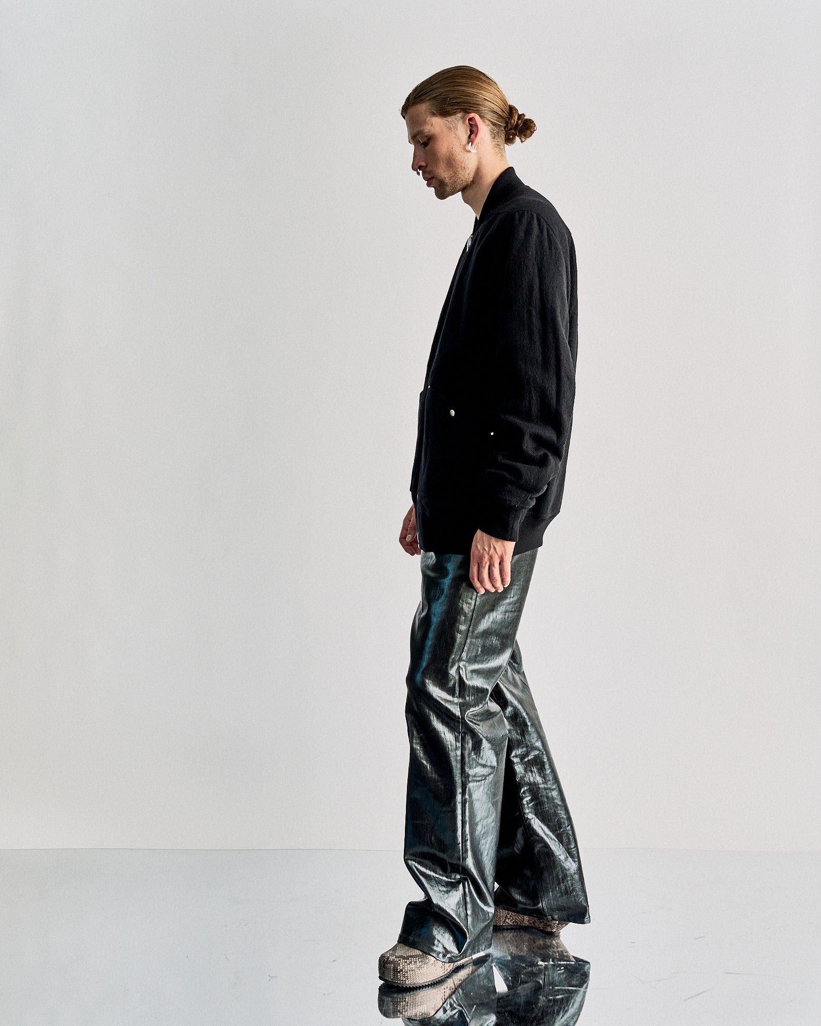 A person with tied-back hair stands sideways, wearing a black Rick Owens bomber jacket and shiny metallic pants. They appear contemplative against a plain backdrop.