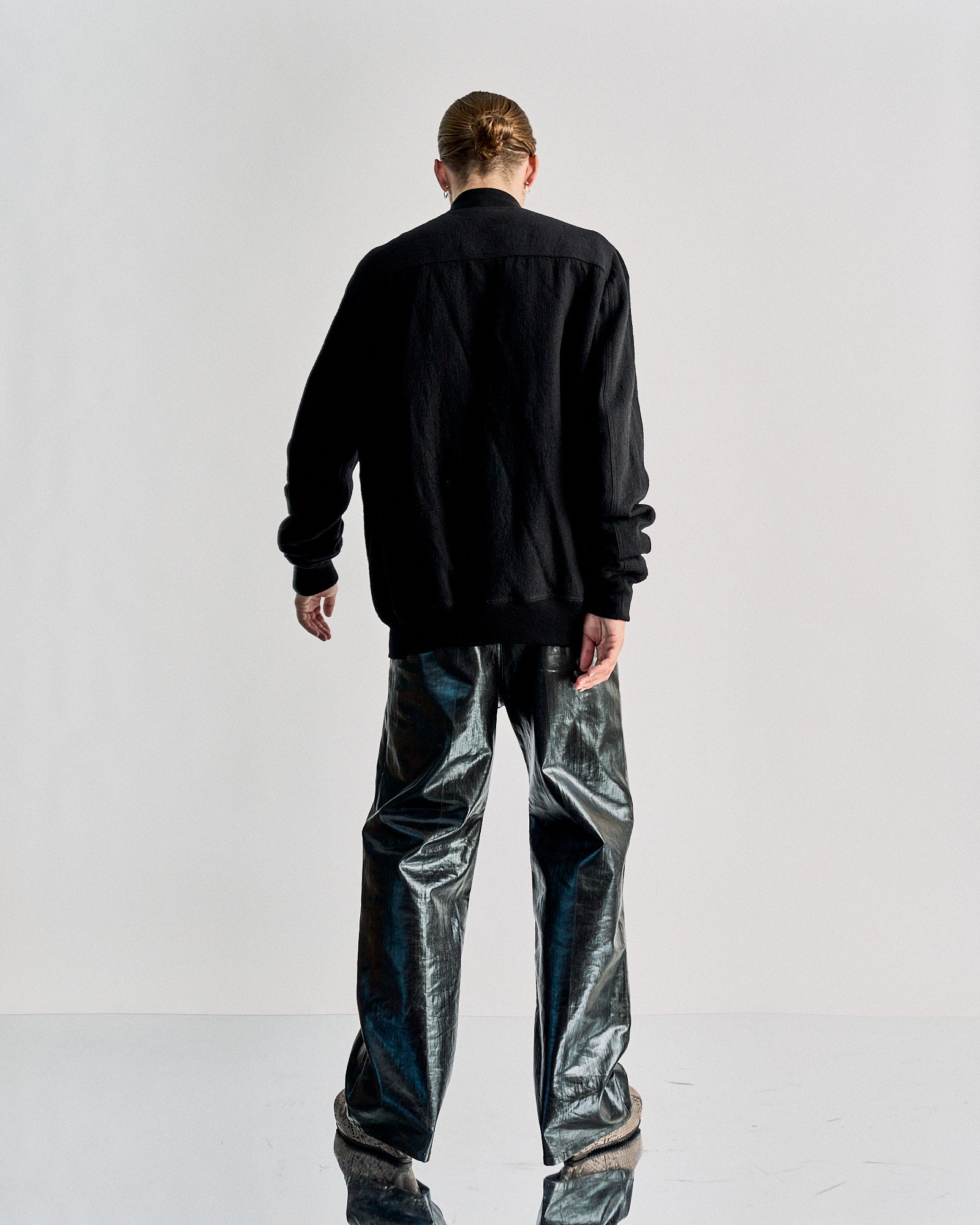 A person stands facing away, wearing a black Rick Owens jacket and shiny metallic pants. Their hair is tied back. The setting is minimalistic with a neutral tone.