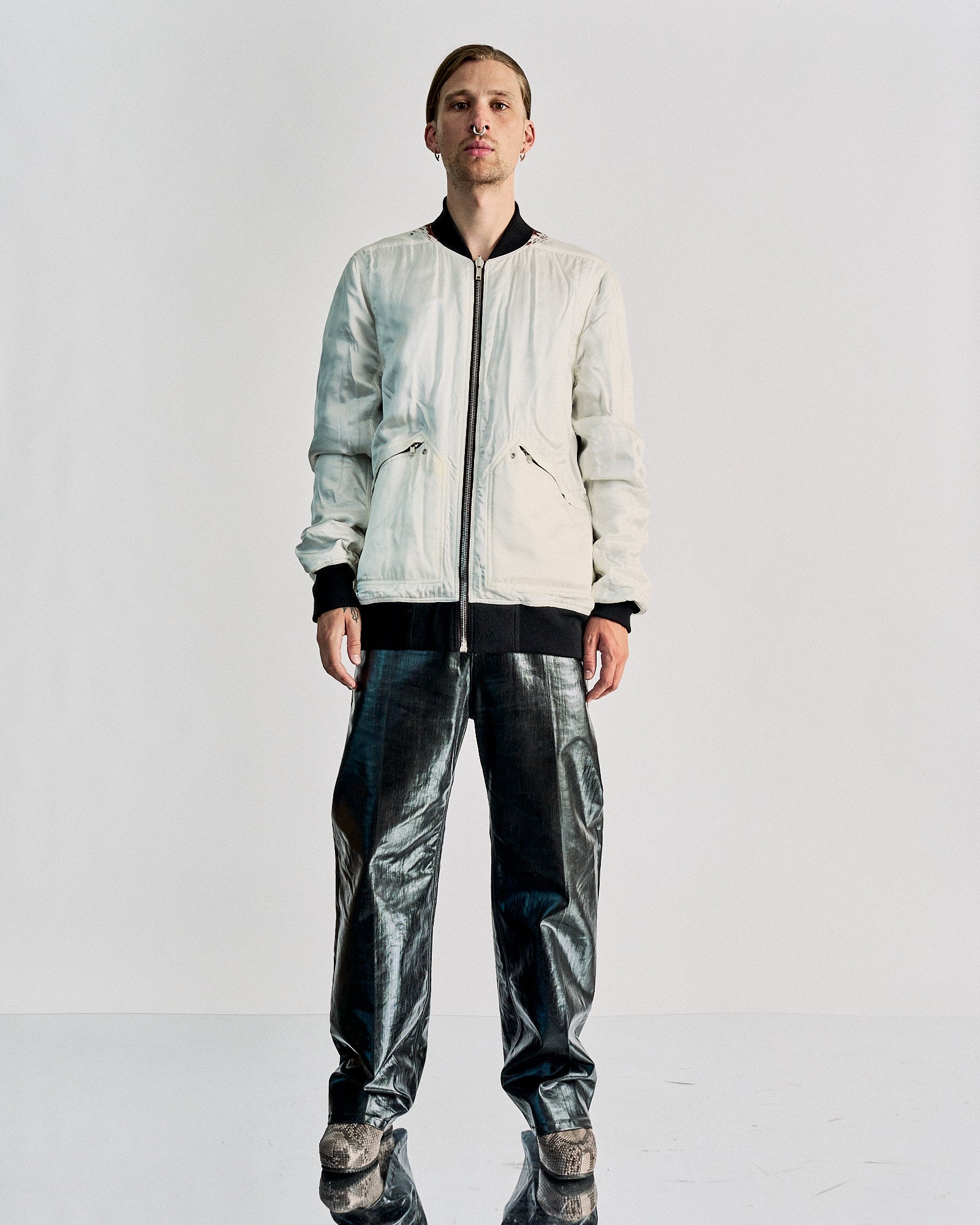 A person stands confidently wearing a white Rick Owens bomber jacket and shiny metallic silver pants. The shoes are patterned, and the background is plain white.
