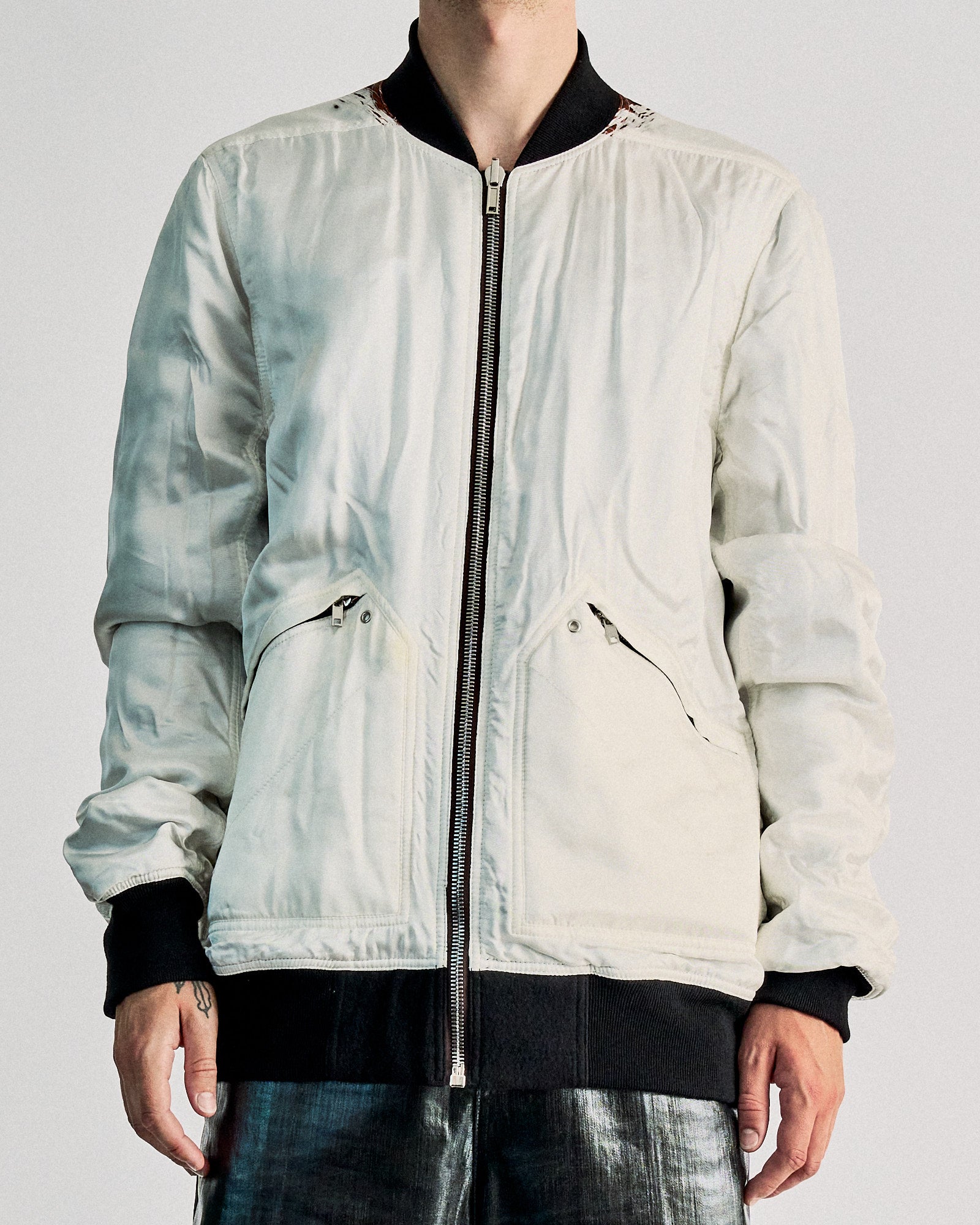 A person stands confidently wearing a white Rick Owens bomber jacket and shiny metallic silver pants. The shoes are patterned, and the background is plain white.