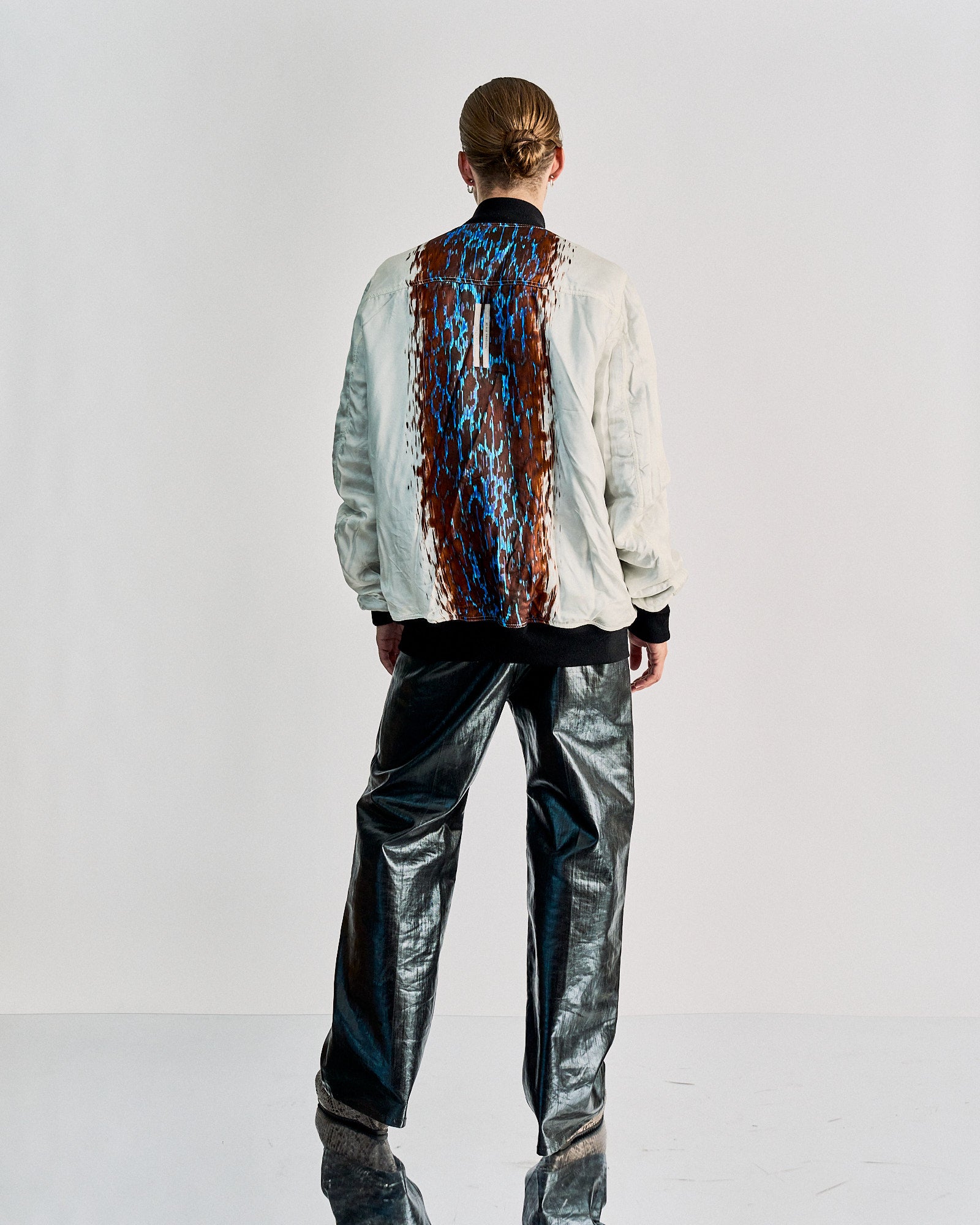 A person with slicked-back hair wearing a white Rick Owens jacket with an abstract blue and brown pattern on the back, paired with shiny black pants.