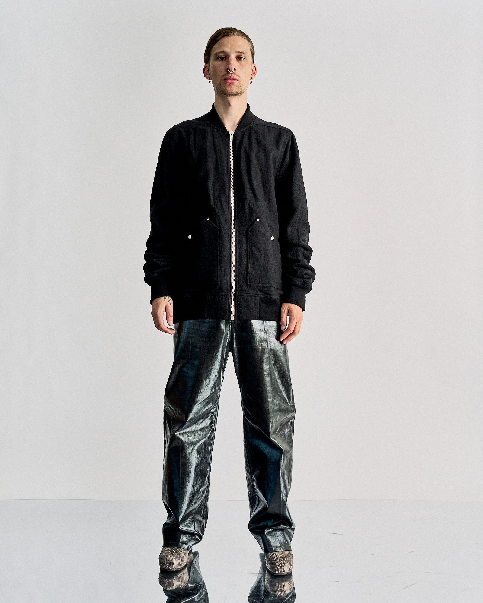 Person wearing a black zip-up Rick Owens jacket and shiny black pants stands on a reflective surface. The minimalist background creates a modern, edgy tone.