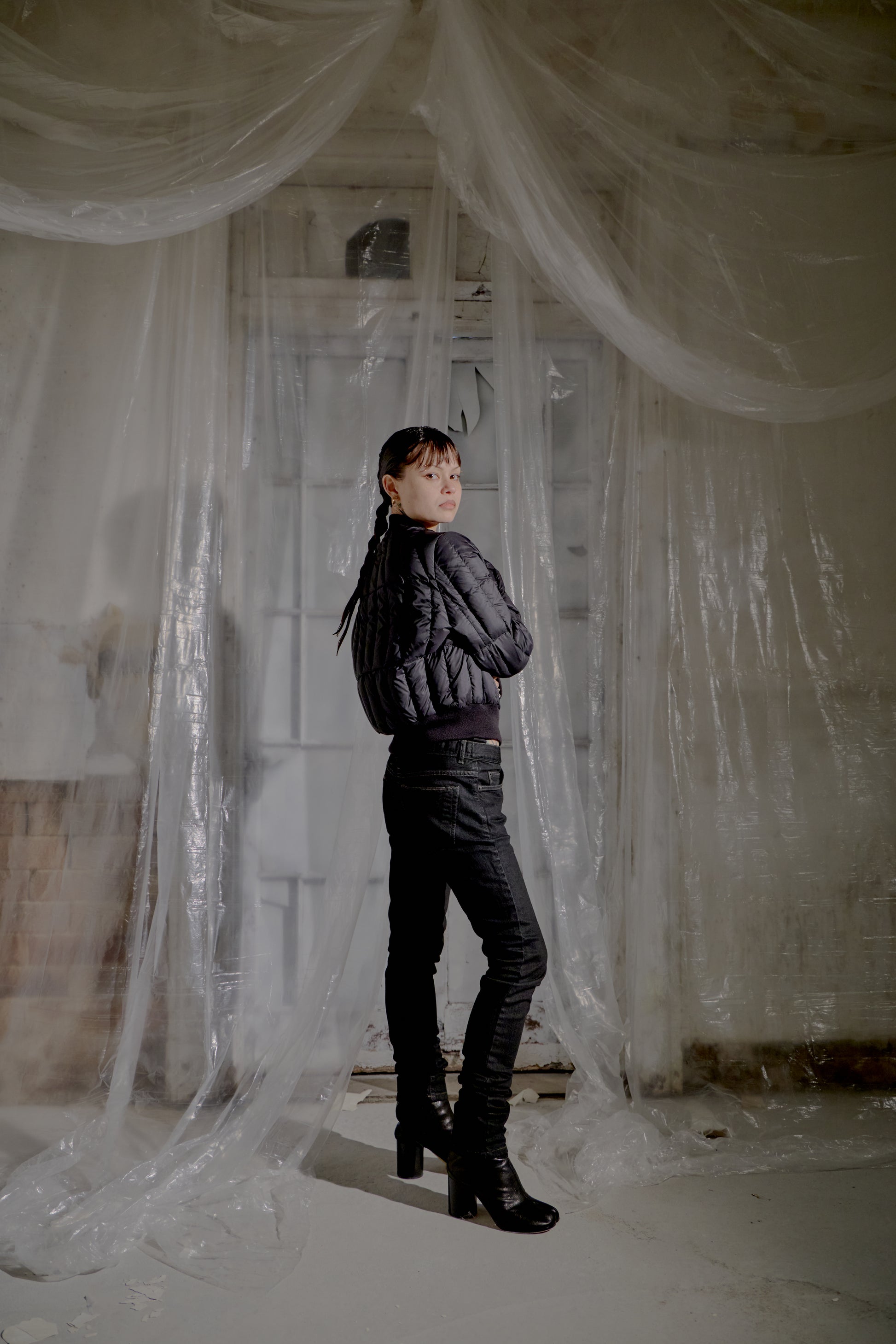 Person standing in a room with plastic sheeting draped over the walls and floor and wearing a Rick Owens black puffer jacket 