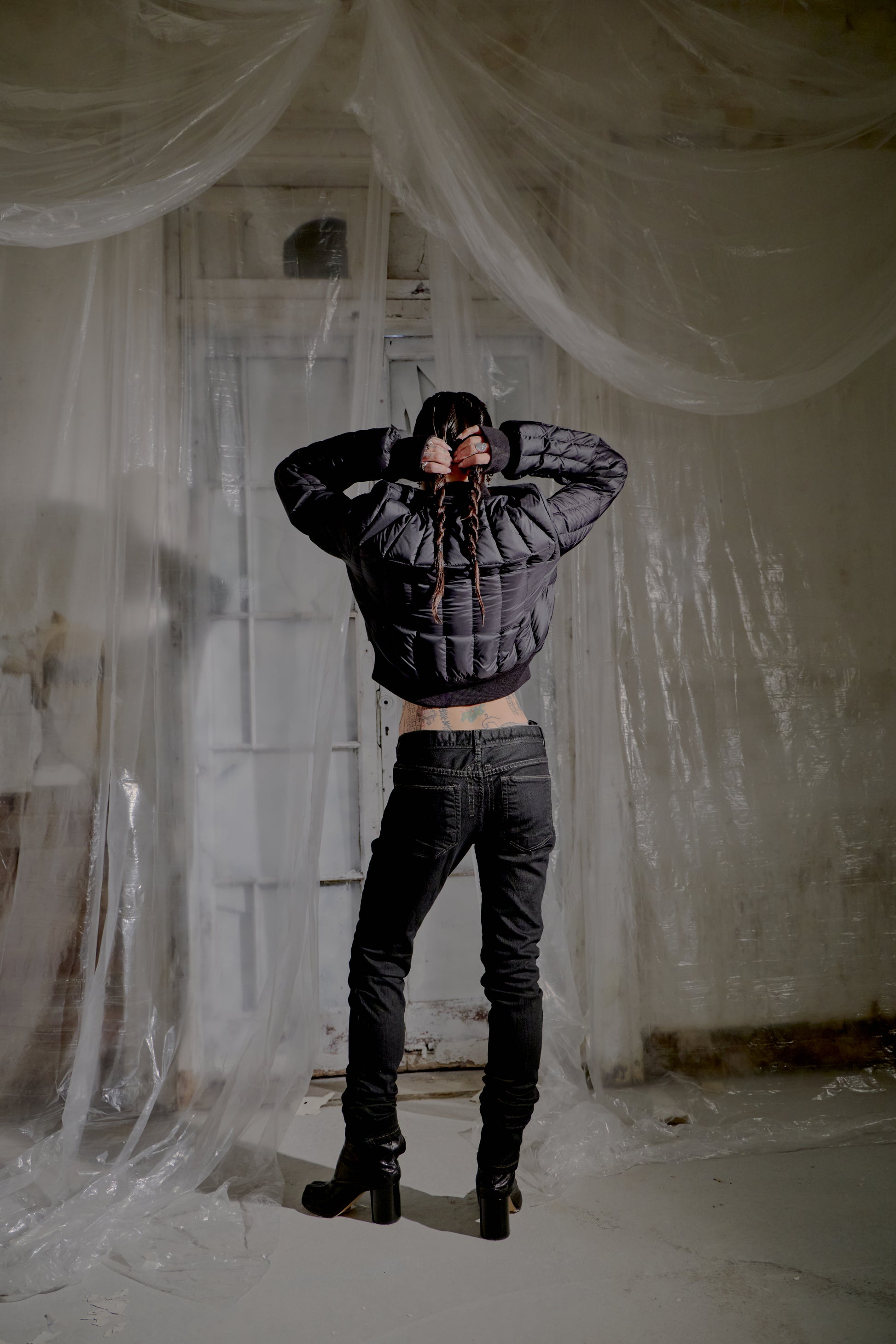 Person wearing a black Rick Owens puffer jacket and dark jeans standing in a room with plastic sheeting on the walls.