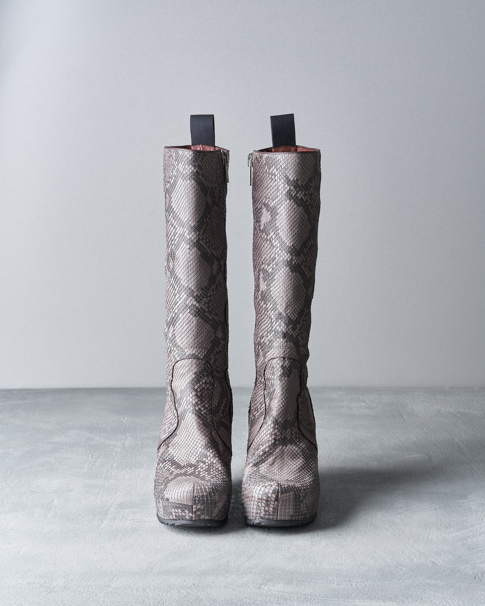 Grey knee-high Rick Owens boots with a snakeskin pattern stand upright on a textured grey surface.