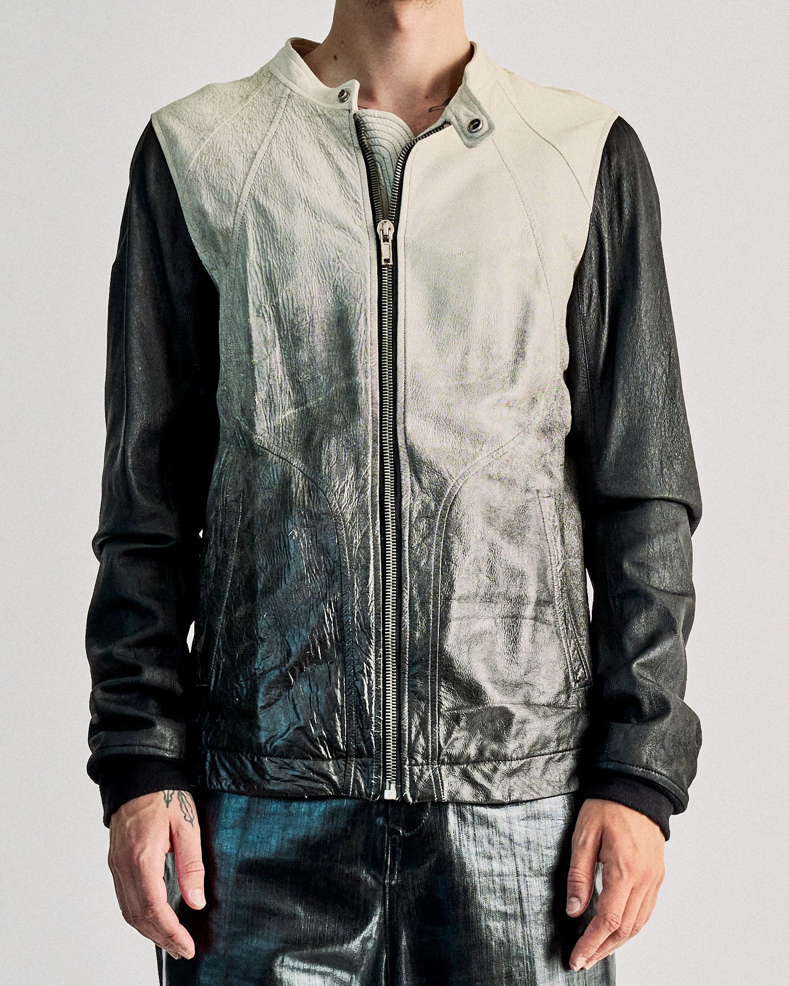 A person wearing a two-toned leather Rick Owens jacket with a white upper half and black sleeves stands against a plain background. The jacket features a central zipper.