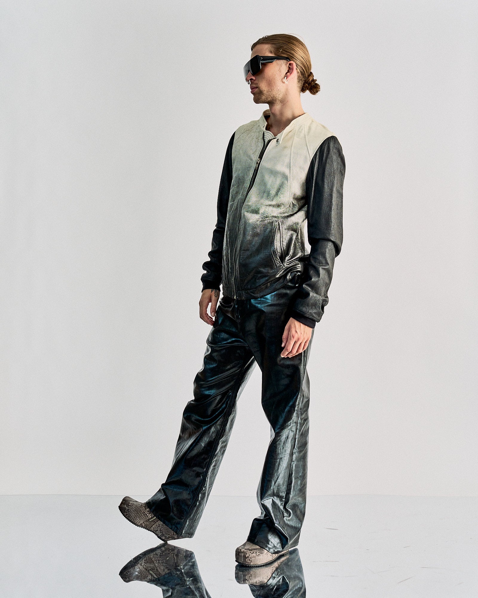 Person with long hair in bun wearing ombre black-and-grey Rick Owens jacket, shiny black pants, and textured shoes. Wears dark sunglasses; cool, futuristic vibe.