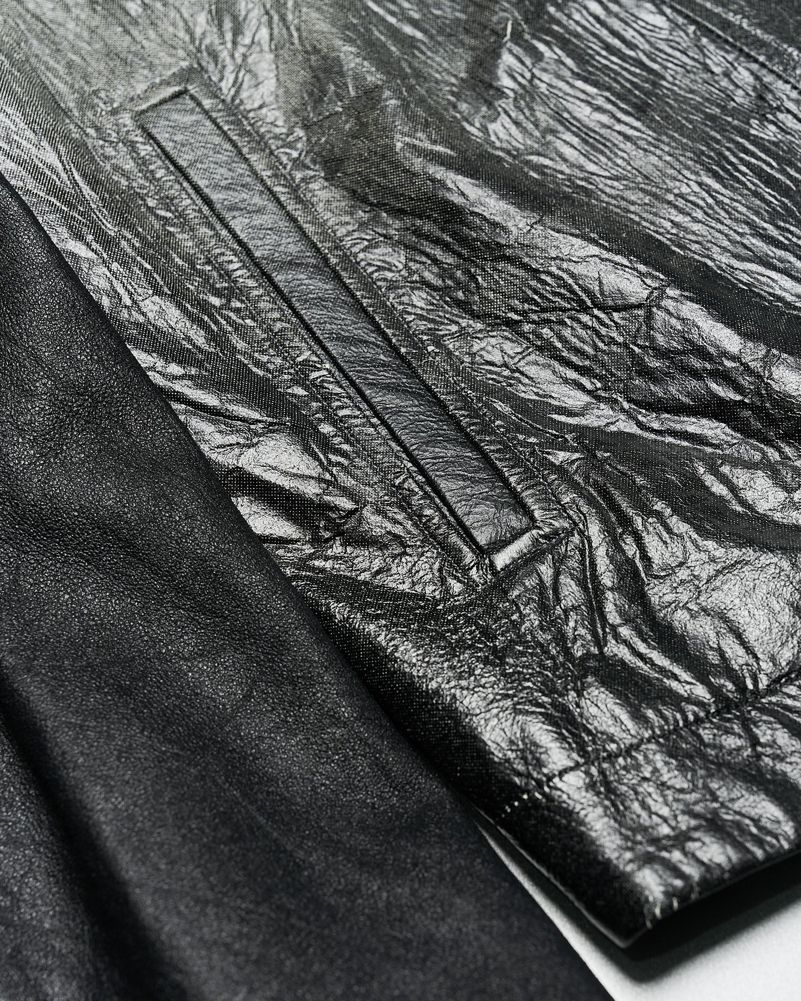 Close-up of a black leather Rick Owens jacket showing contrasting textures. The left side is smooth, while the right side has a crinkled appearance, with a visible pocket seam.