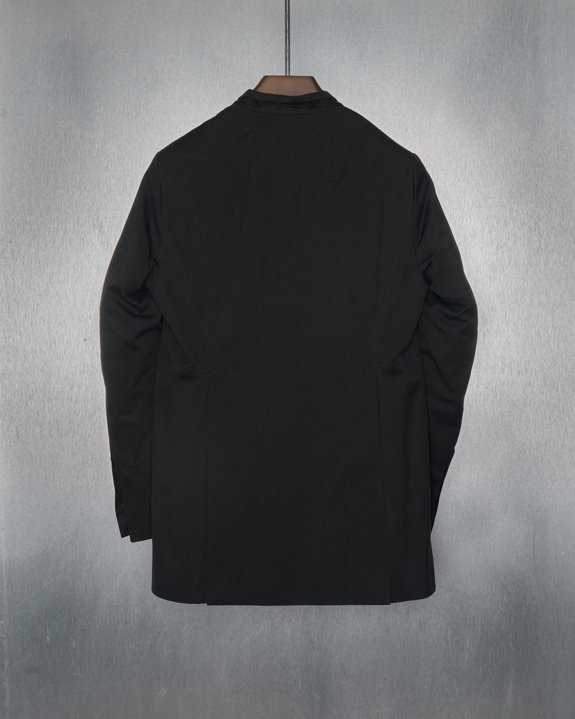 The reverse of a black Rick Owens blazer with a minimalist half-collar, grid panelled construction, and a leather single-button closure. The jacket is displayed against a textured grey background.