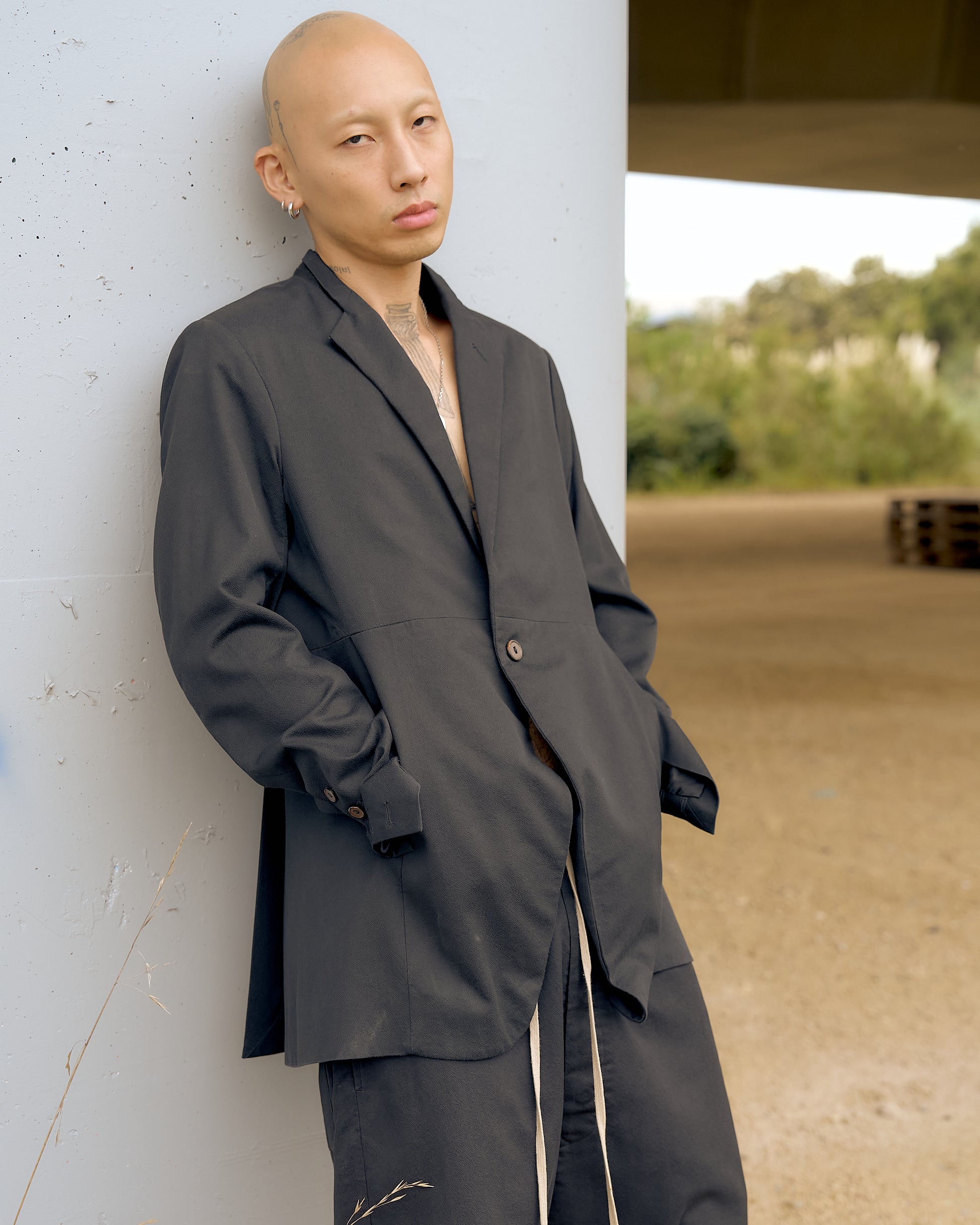 Bald person leaning against a pillar, wearing a loose-fitting Rick Owens black suit, with a neutral expression. Background features blurred greenery.