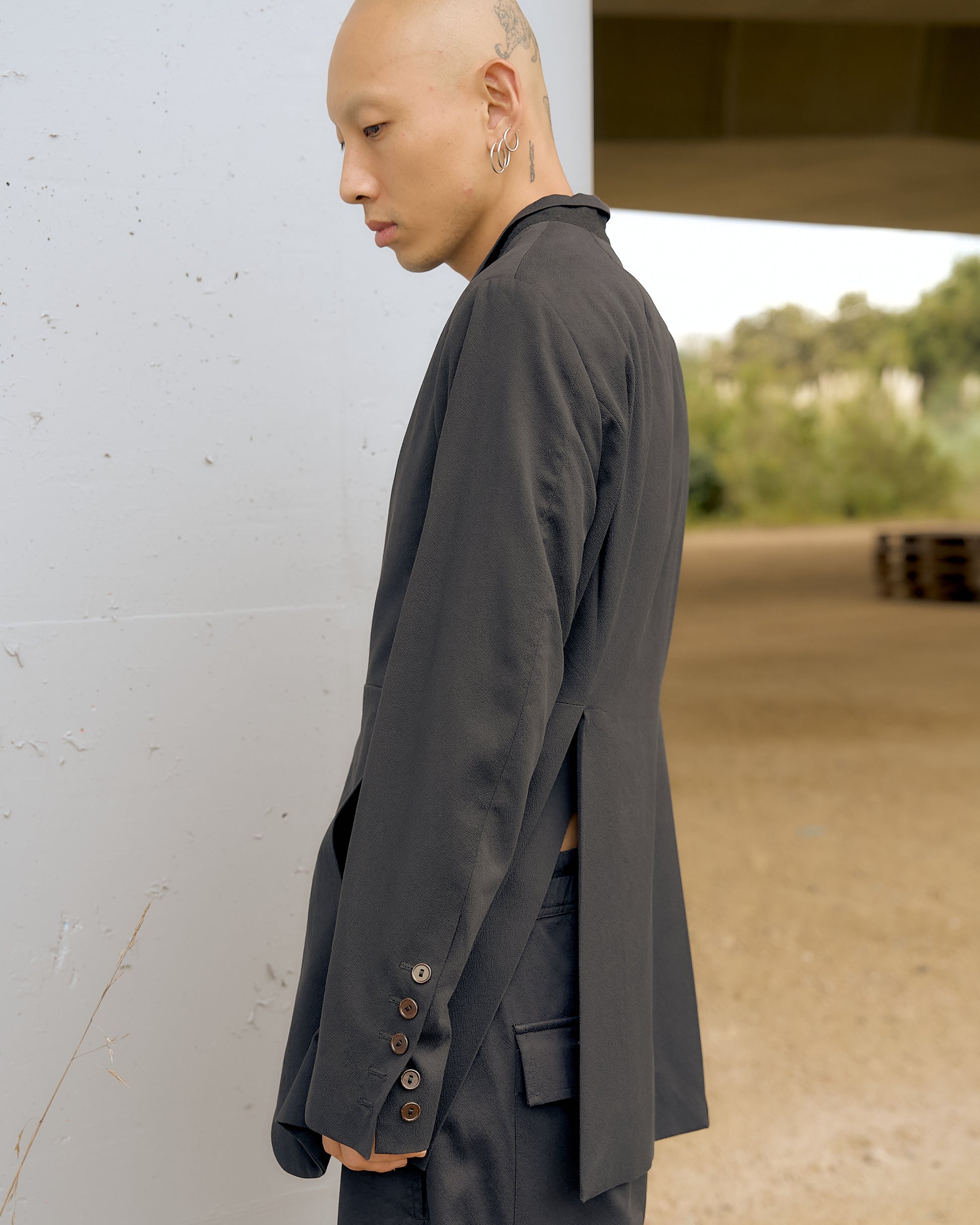 Bald person in a stylish, dark, open-back Rick Owens suit with button details stands by a light-coloured wall. The background has a neutral, outdoor setting.