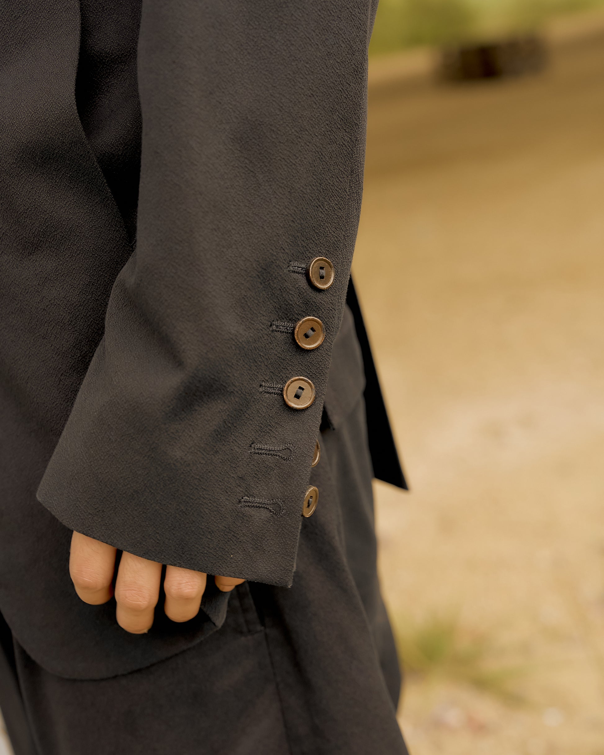 Close-up of a person in a dark suit, showing the sleeve with brown buttons and a relaxed hand by their side. Background is a soft, blurred outdoor scene.