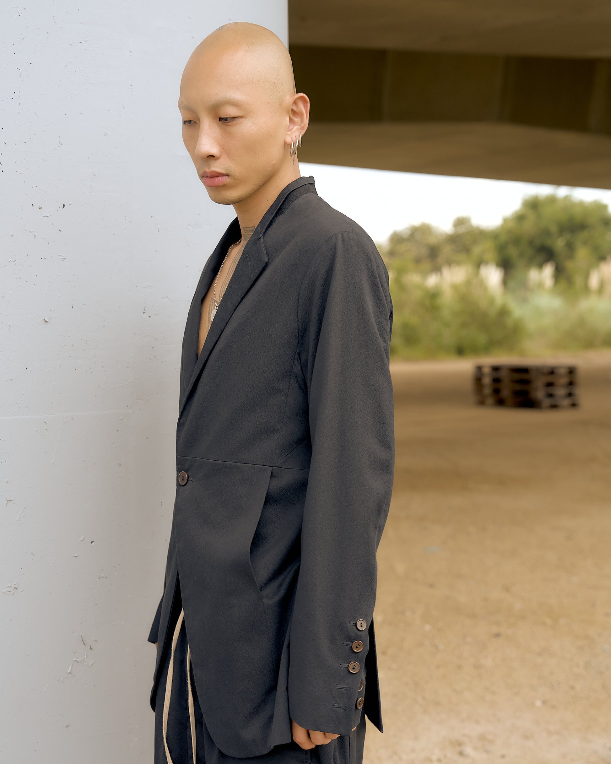 A person with a bald head and earrings stands pensively in a dark Rick Owens blazer, leaning against a white column. Open space and greenery are in the background.