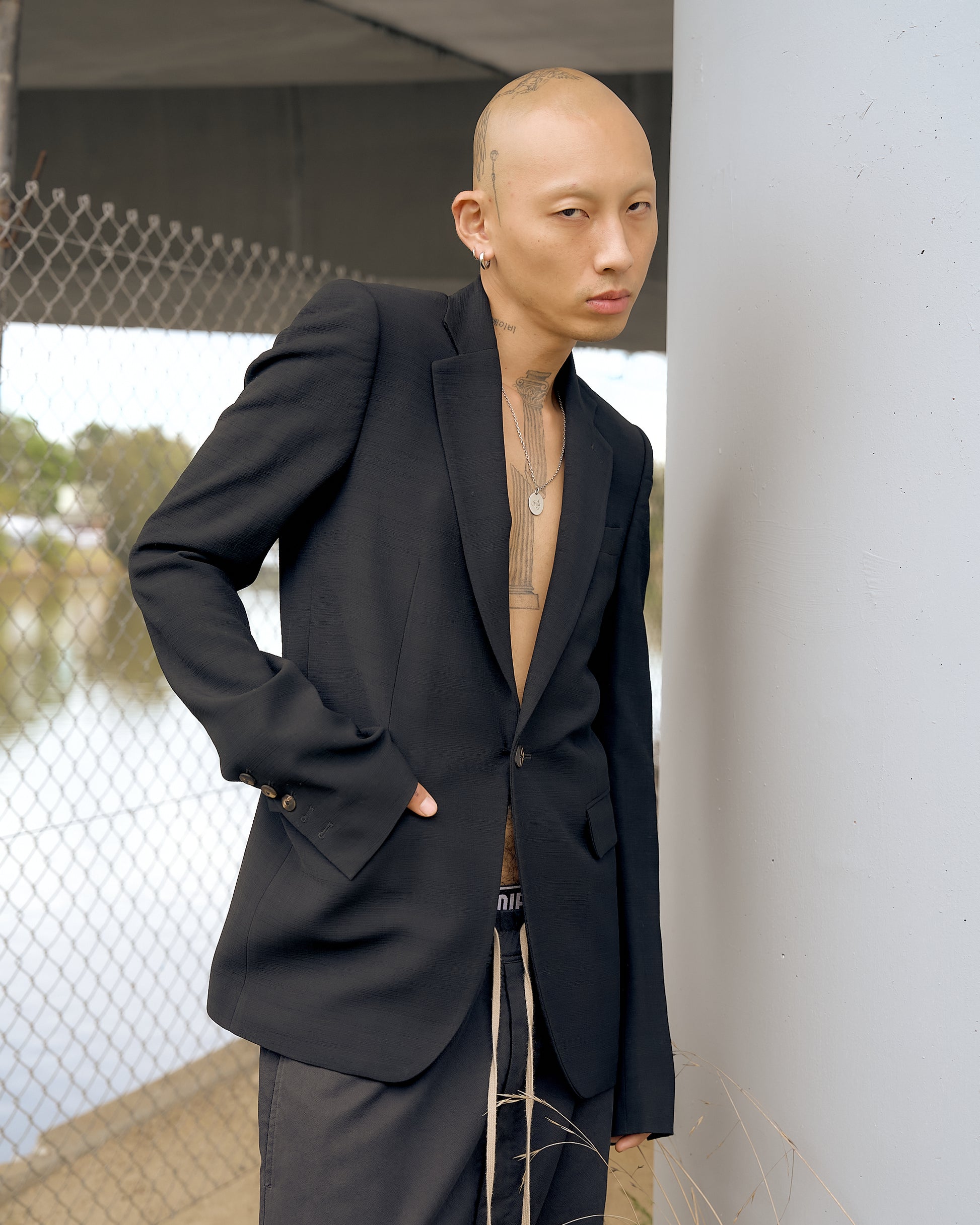 A person with a bald head and tattoos wears a black Rick Owens blazer over loose pants, standing confidently by a chain-link fence and concrete pillar.