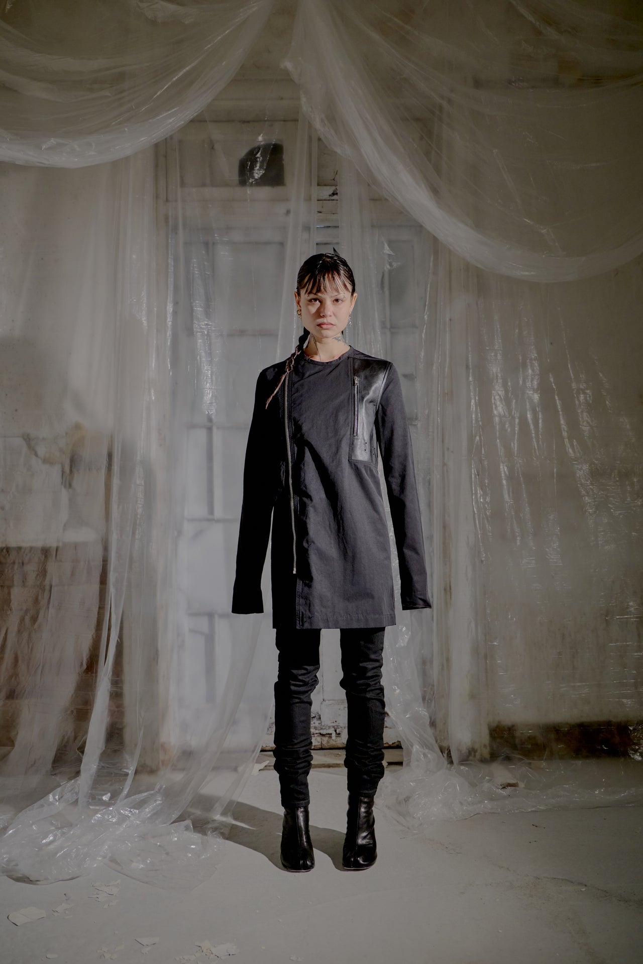 Person wearing a dark Rick Owens coat and boots standing in a room with white fabric draped around.