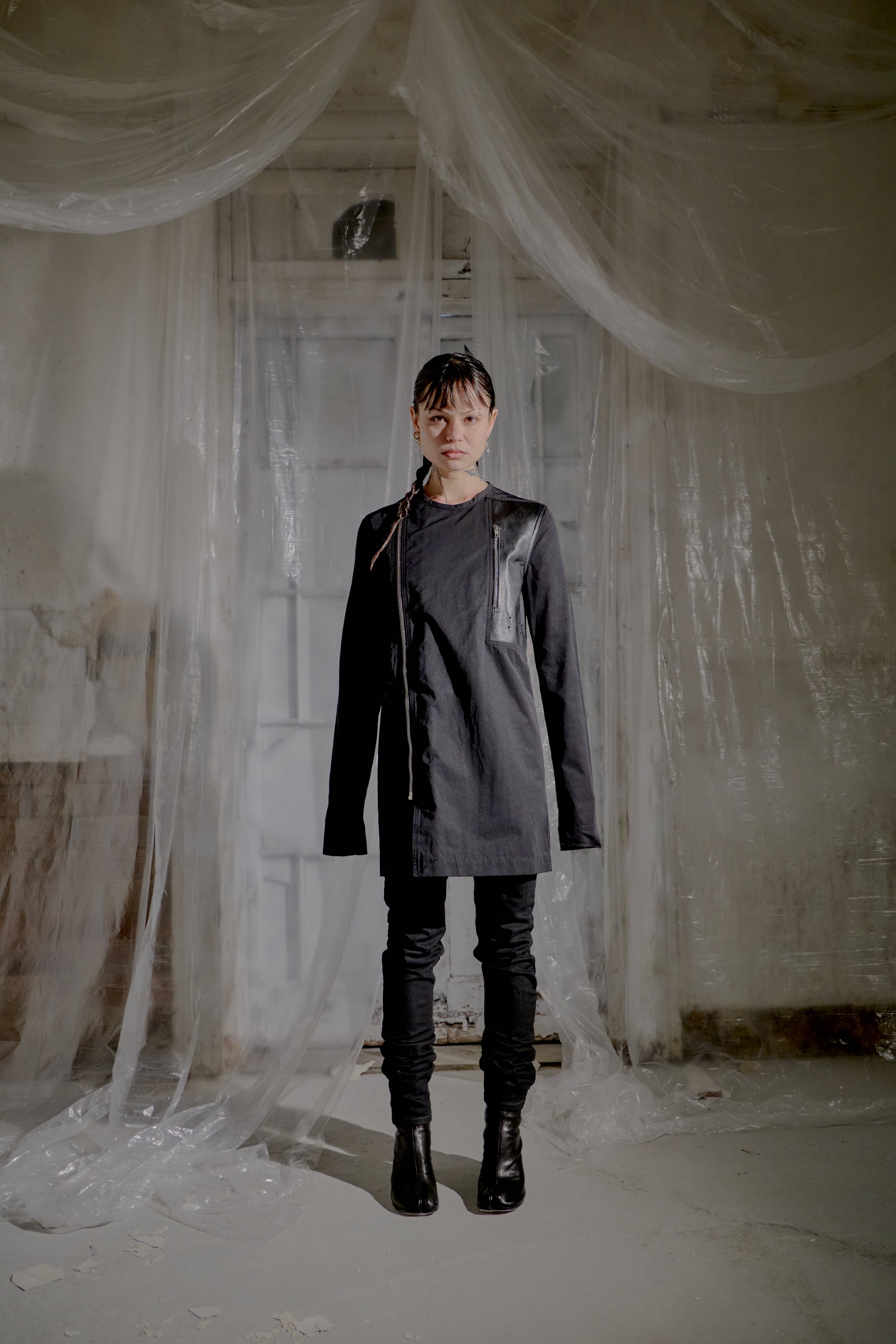 Person wearing a dark Rick Owens coat and boots standing in a room with white fabric draped around.