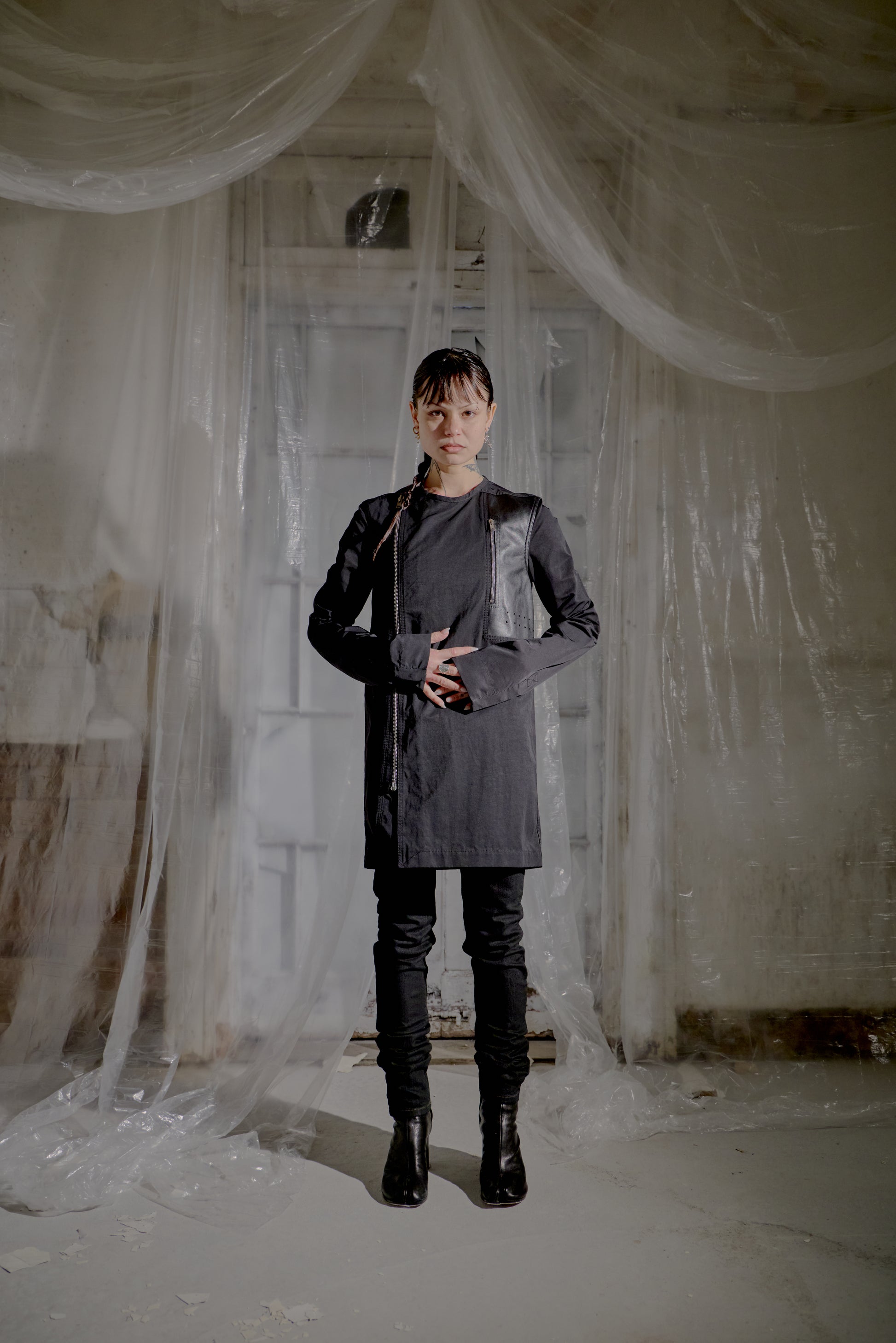 Person wearing a long dark Rick Owens coat and black pants standing in a room with white draped fabric.