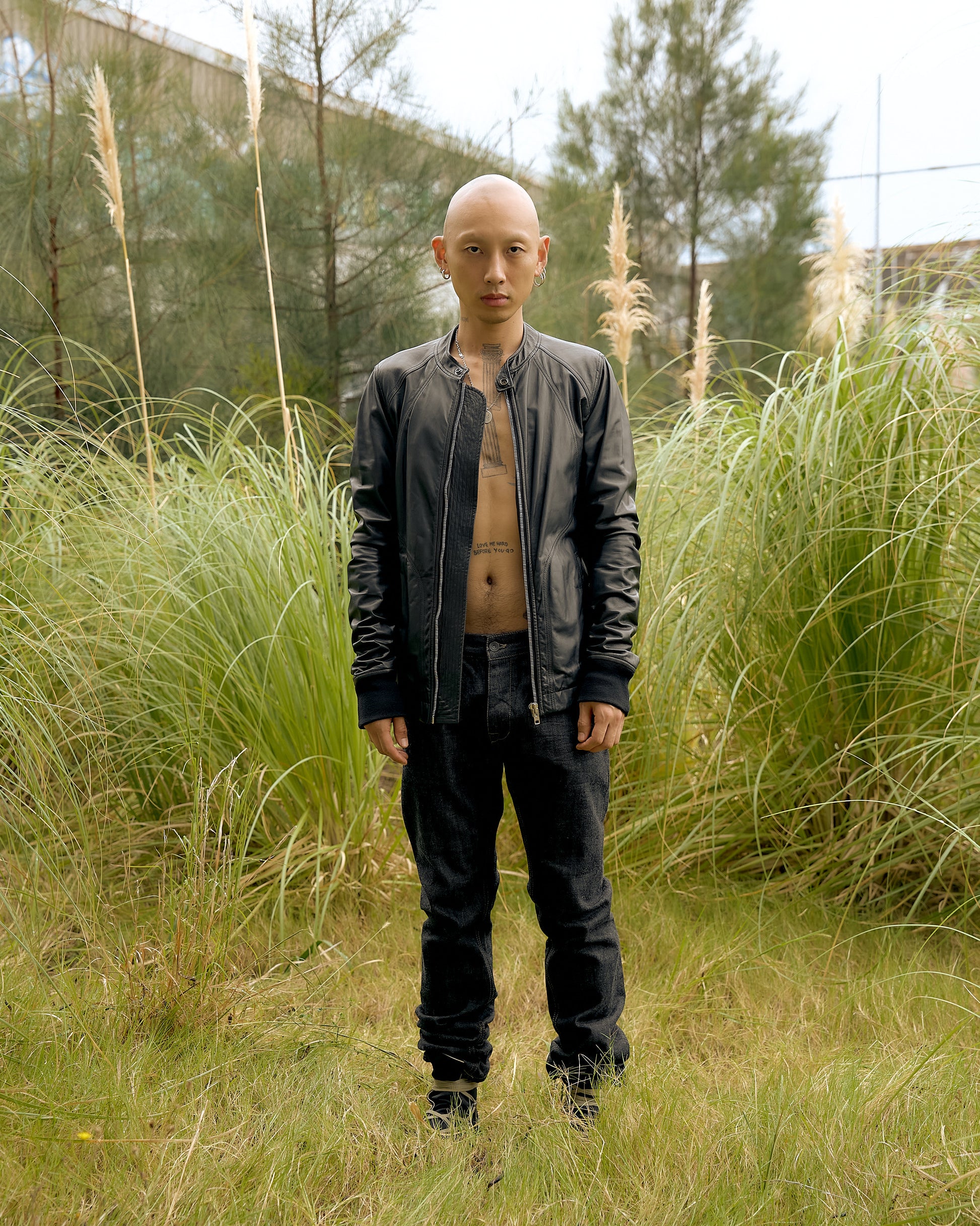 A person wearing a black Rick Owens leather jacket with a mandarin collar, standing in a natural setting with tall grass and trees in the background.