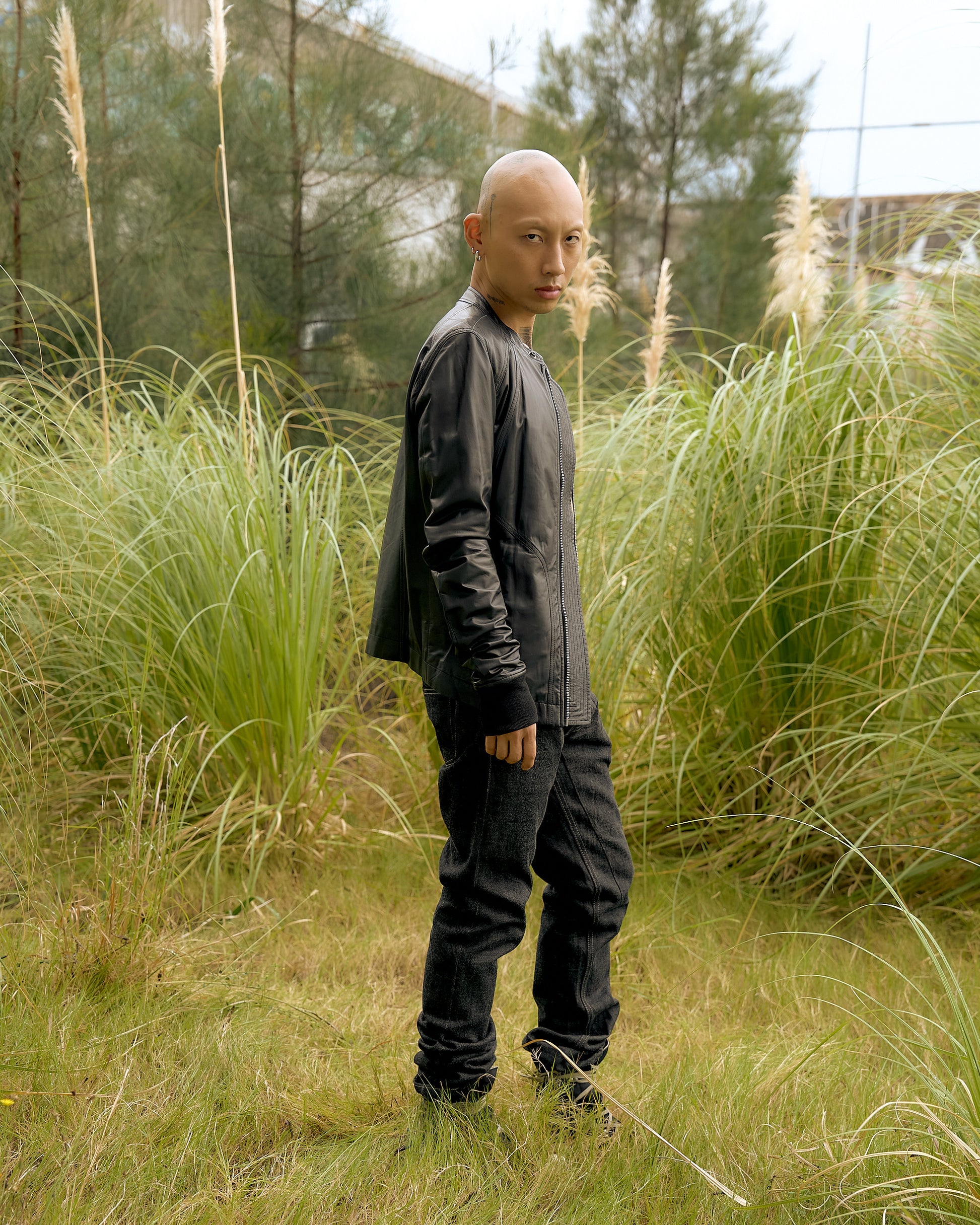 A person in a black leather Rick Owens jacket stands confidently in a grassy field with tall greenery and trees. 