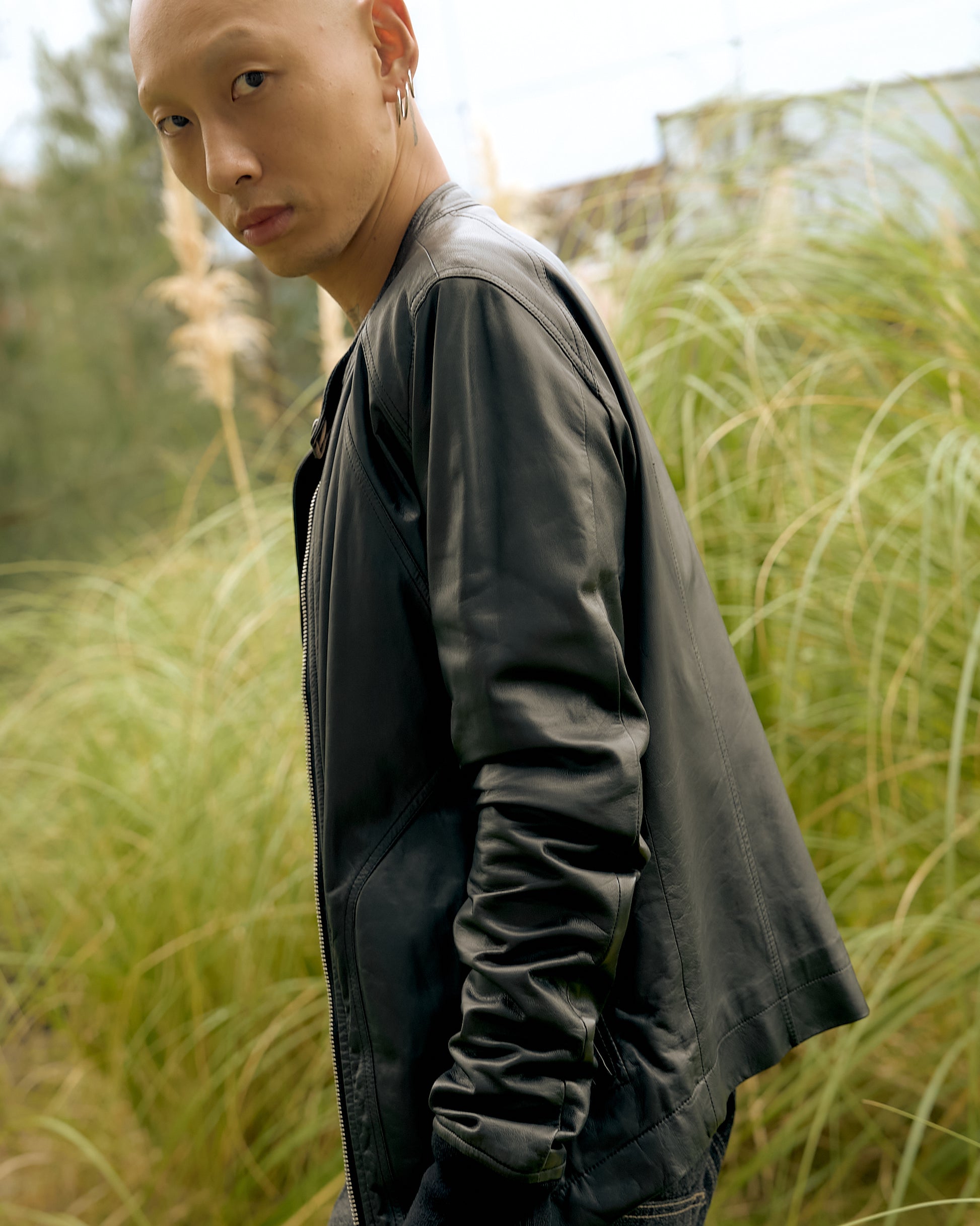 A person with a serious expression wears a black leather Rick Owens jacket, standing in tall grass. 