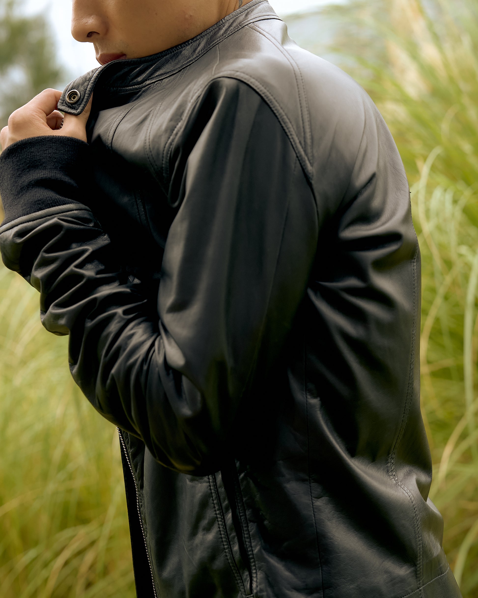 A person in a black leather Rick Owens jacket stands among tall grasses, holding the collar close.